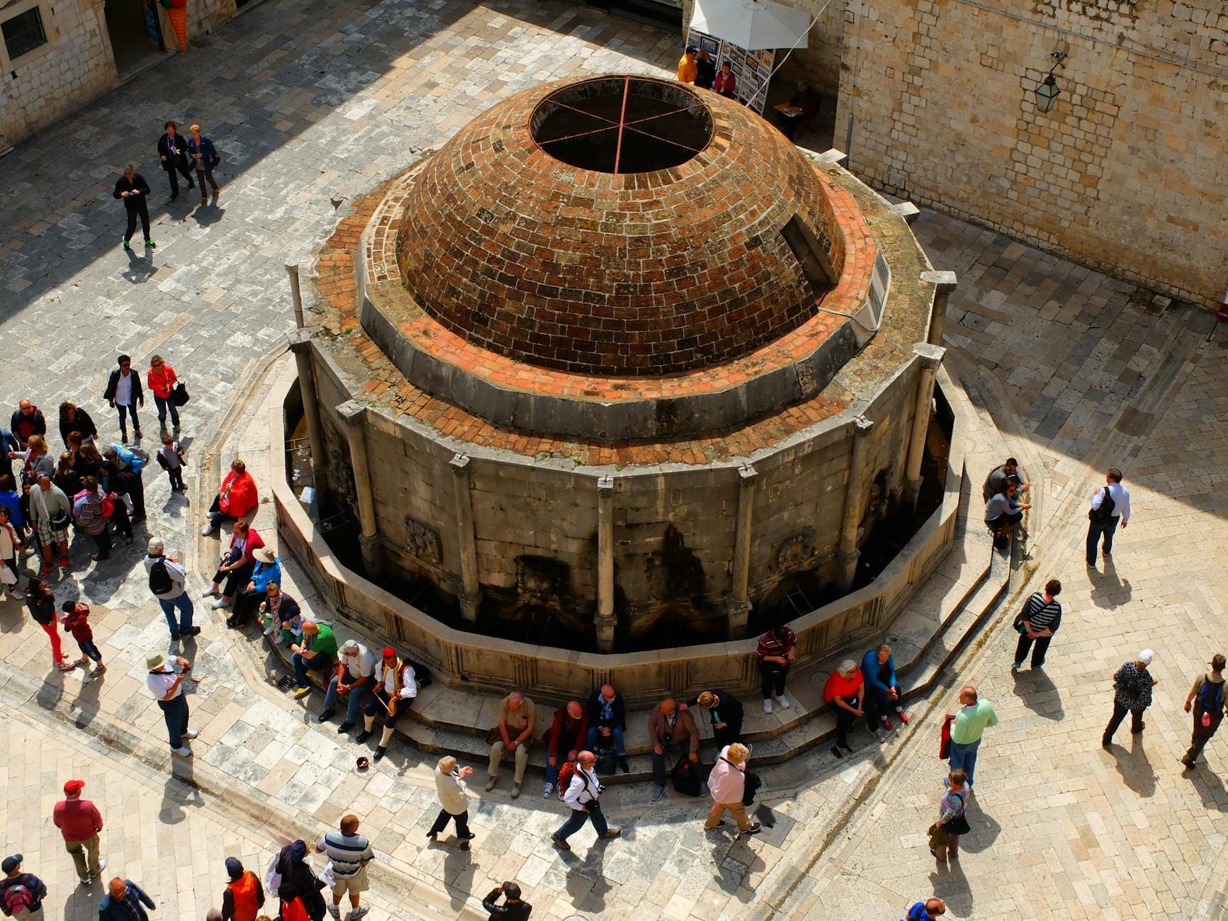 Vista aérea de la Gran Fuente de Onofrio en Dubrovnik, con personas sentadas y caminando alrededor de su estructura circular de piedra.