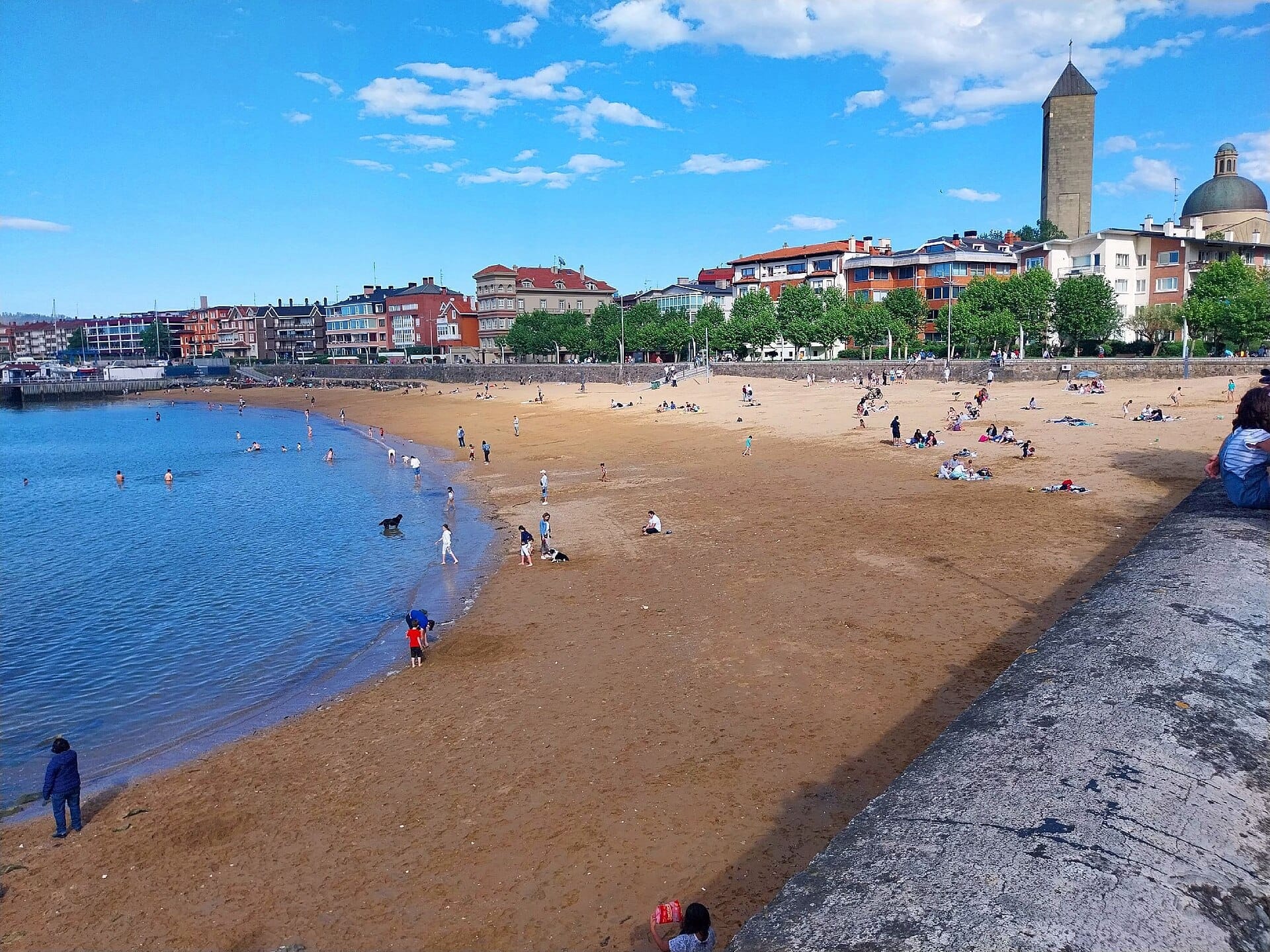 Familien und Besucher genießen den goldenen Sand und das ruhige Wasser am Playa de Las Arenas mit bunten Gebäuden und ikonischen Türmen im Hintergrund.