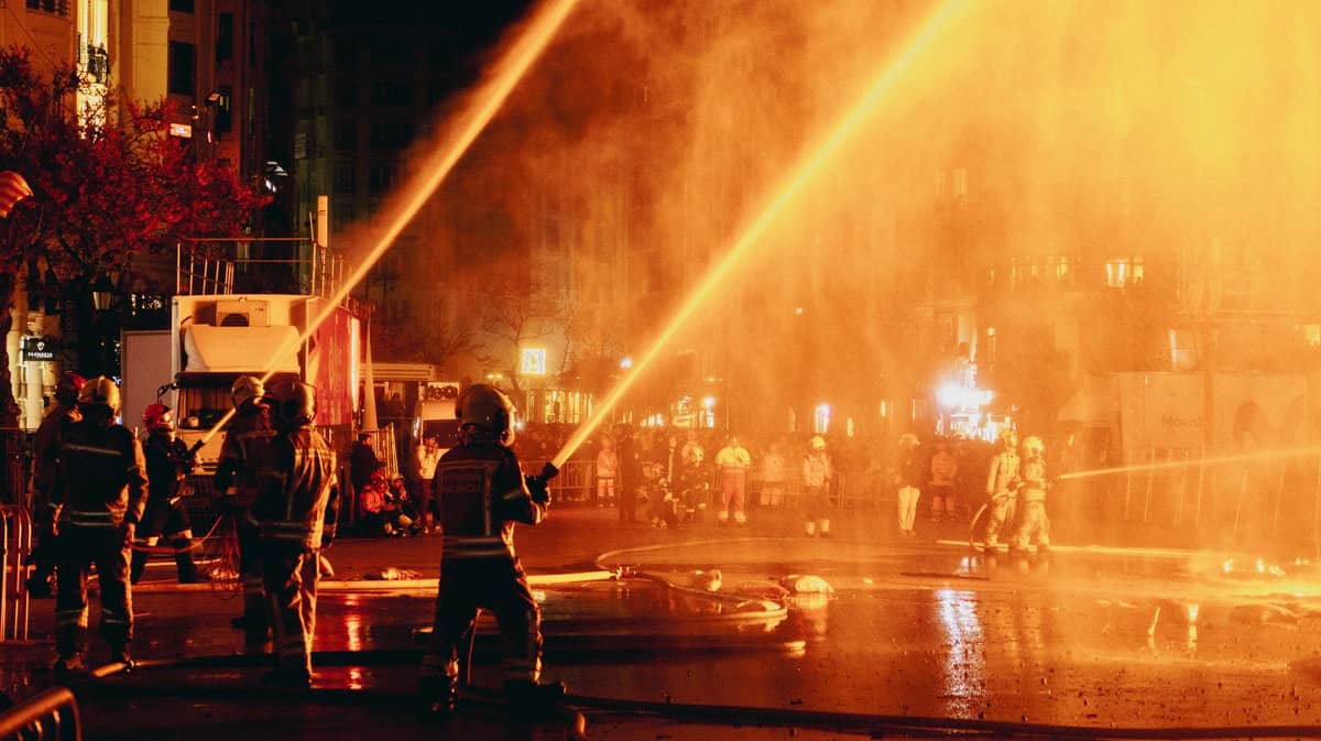 Feuerwehrleute spritzen Wasser inmitten von Rauch und Flammen auf einem Stadtplatz während der Fallas von Valencia bei Nacht