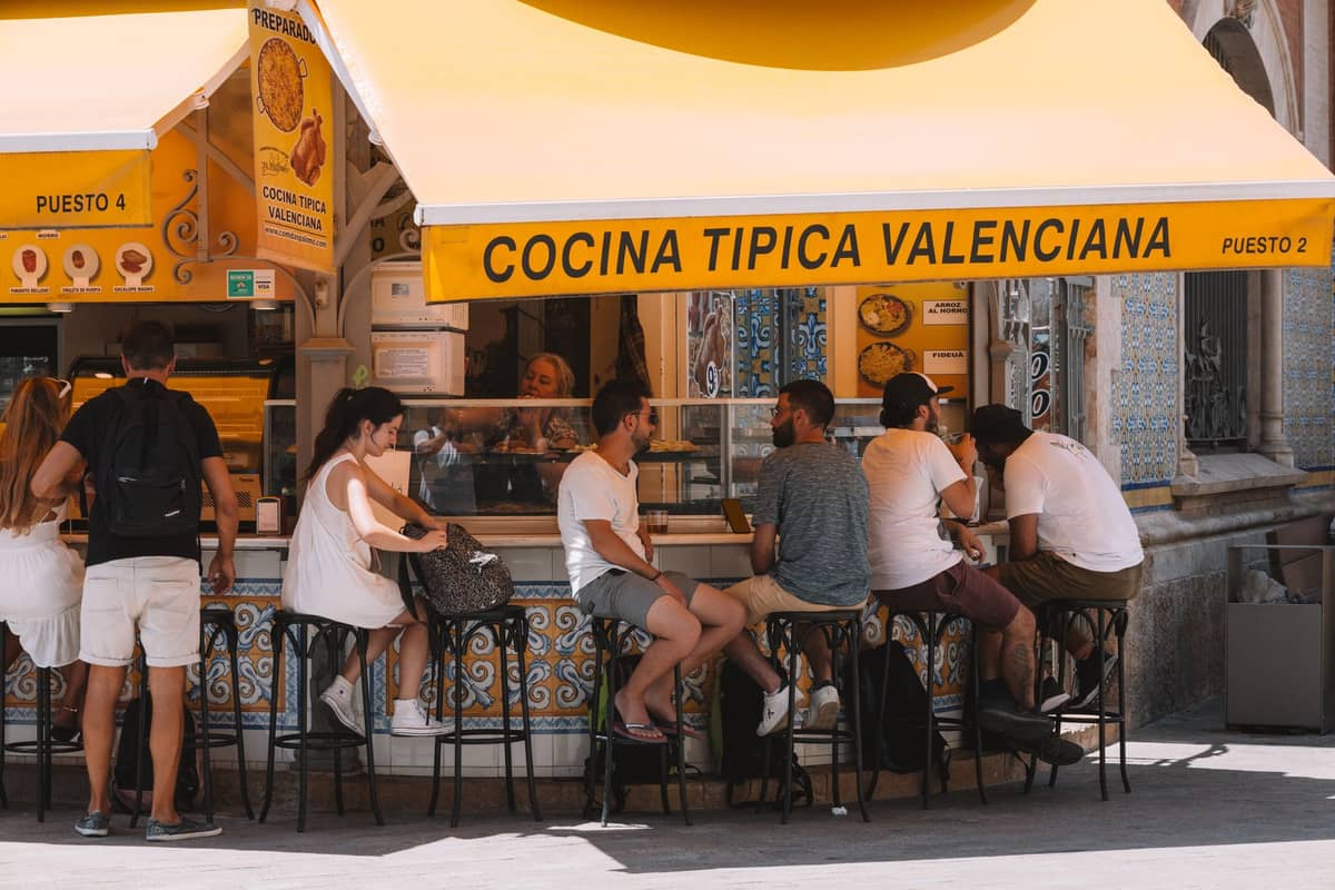 Menschen, die an einem Essensstand mit einem gelben Sonnendach mit der Aufschrift 'Cocina Tipica Valenciana' in Valencia, Spanien, sitzen und essen.