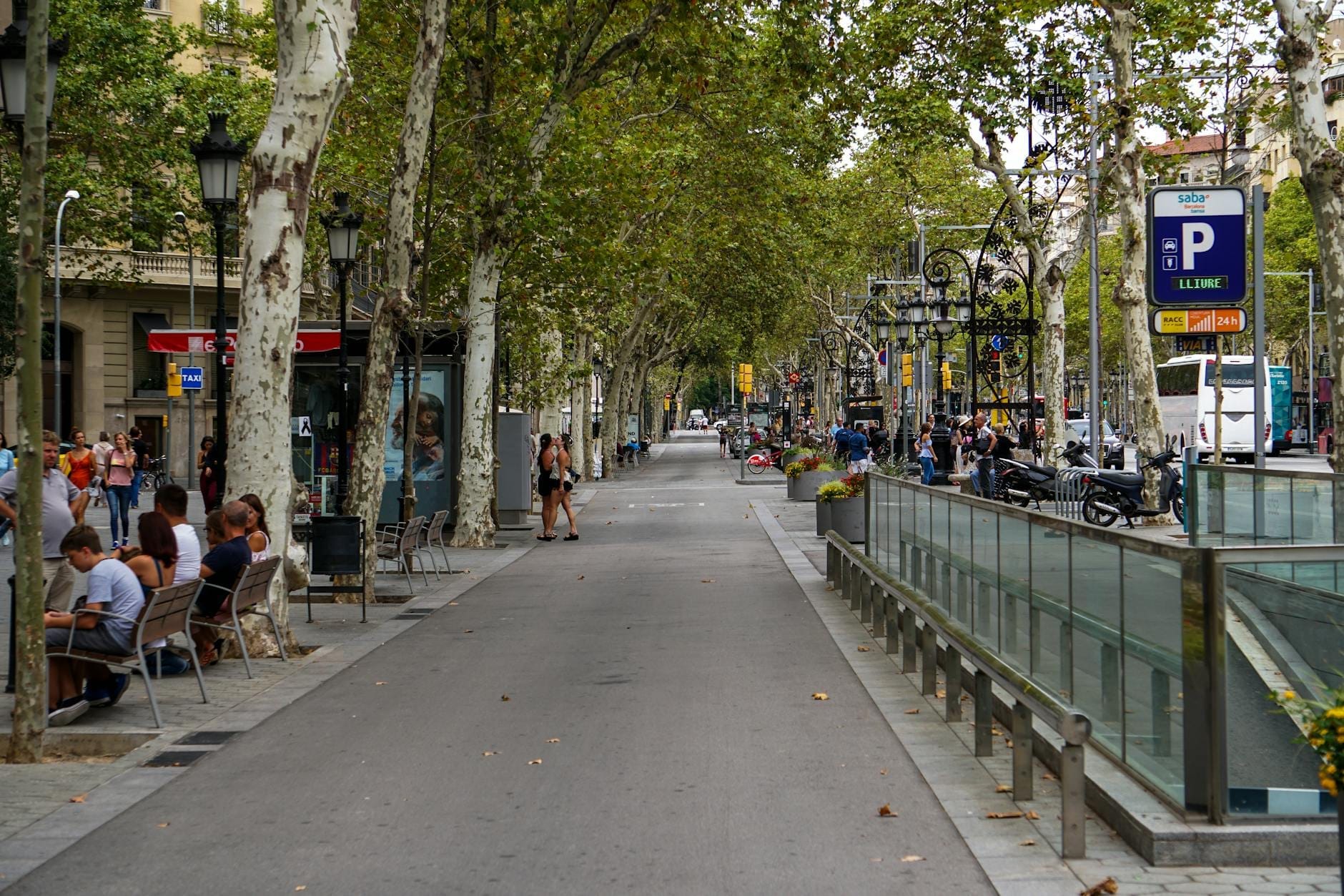 Tree-lined boulevard of Las Ramblas in Barcelona with people sitting at outdoor cafes and pedestrians walking along the wide promenade.