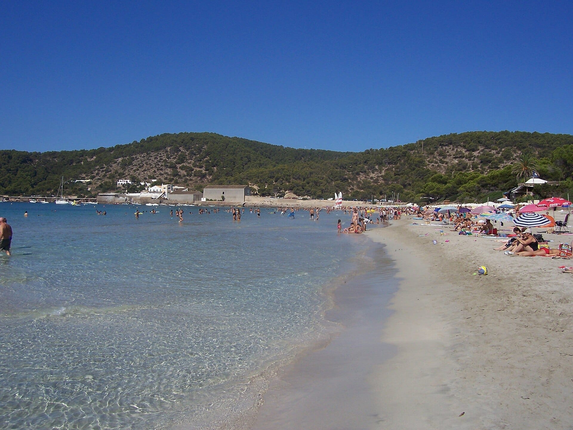 La folla si rilassa sotto ombrelloni colorati lungo la riva limpida della Platja de Ses Salines, con colline coperte di pini ed edifici bianchi sullo sfondo.