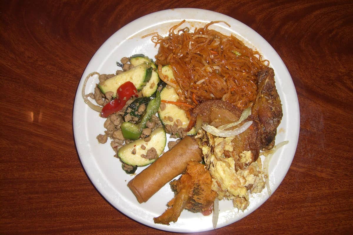 A plate filled with a variety of buffet foods, including noodles, stir-fried vegetables, egg, spring roll, and fried chicken, on a wooden table.