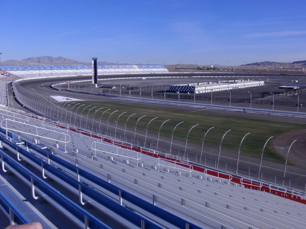 View of the curved track, grandstands, and infield buildings at Las Vegas Motor Speedway under a clear blue sky.