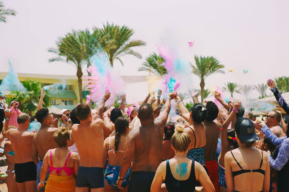 Menschenmenge in Badekleidung bei einer Outdoor-Poolparty mit Palmen, blauem Himmel und buntem Pulver in der Luft bei sonnigem Wetter.