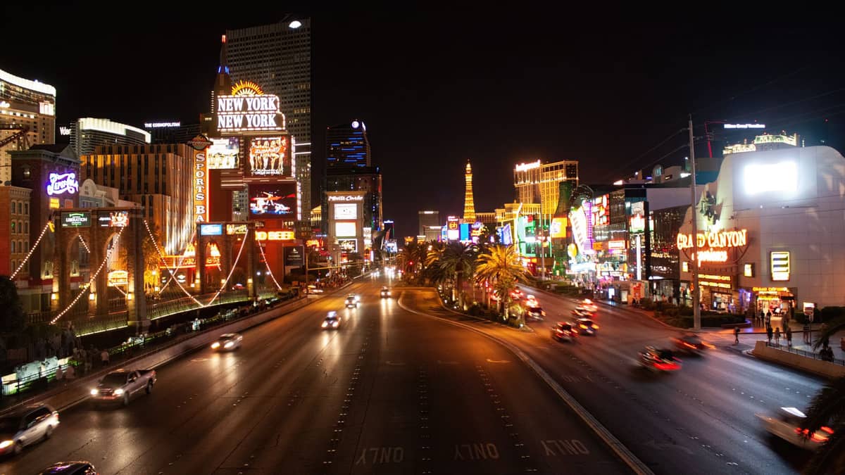 Escena nocturna del Strip de Las Vegas con brillantes luces de neón, el hotel New York New York, otros casinos temáticos y coches circulando.