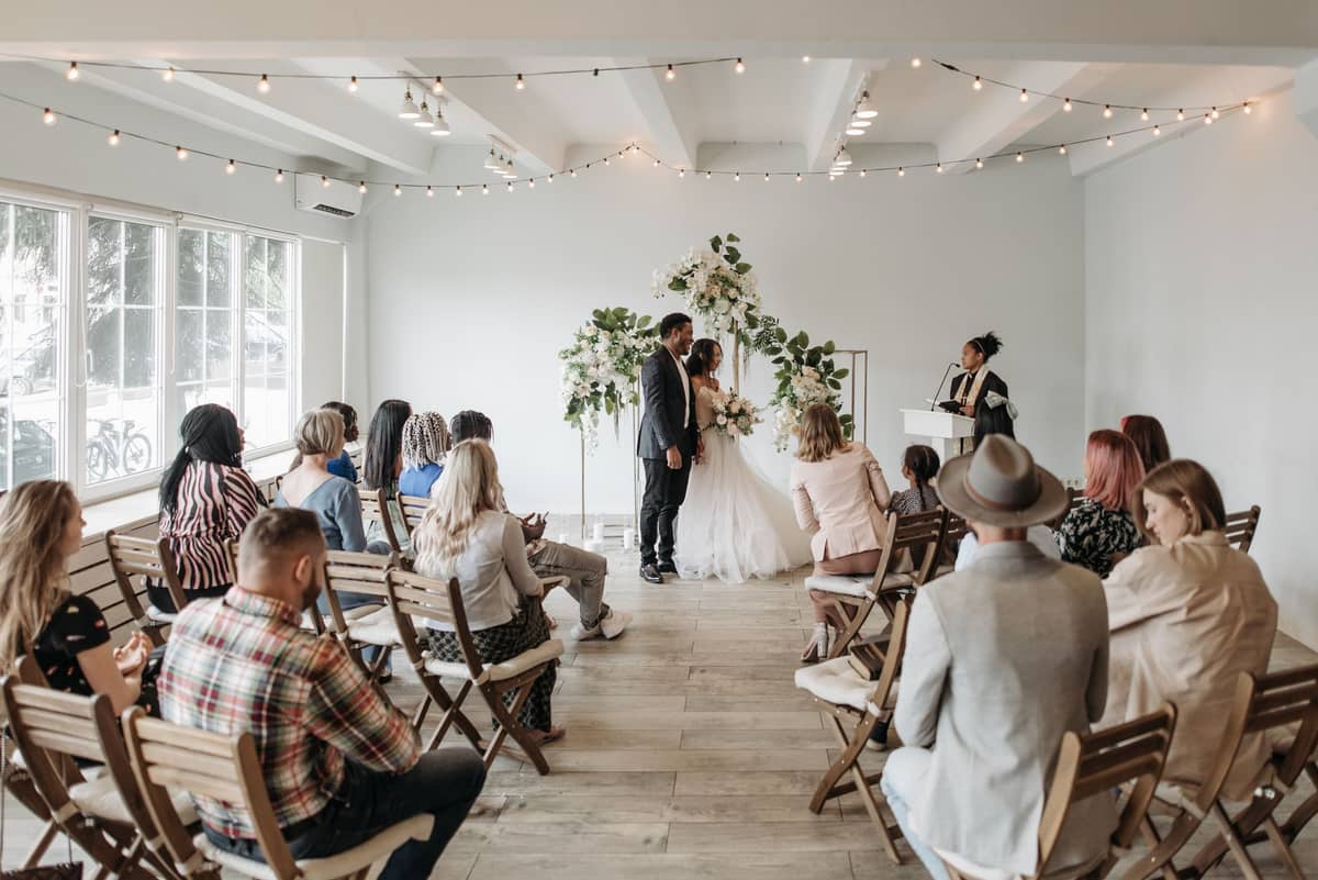 Une cérémonie de mariage dans une petite chapelle moderne, avec des invités assis et un couple devant l'officiant, sous des décorations florales.