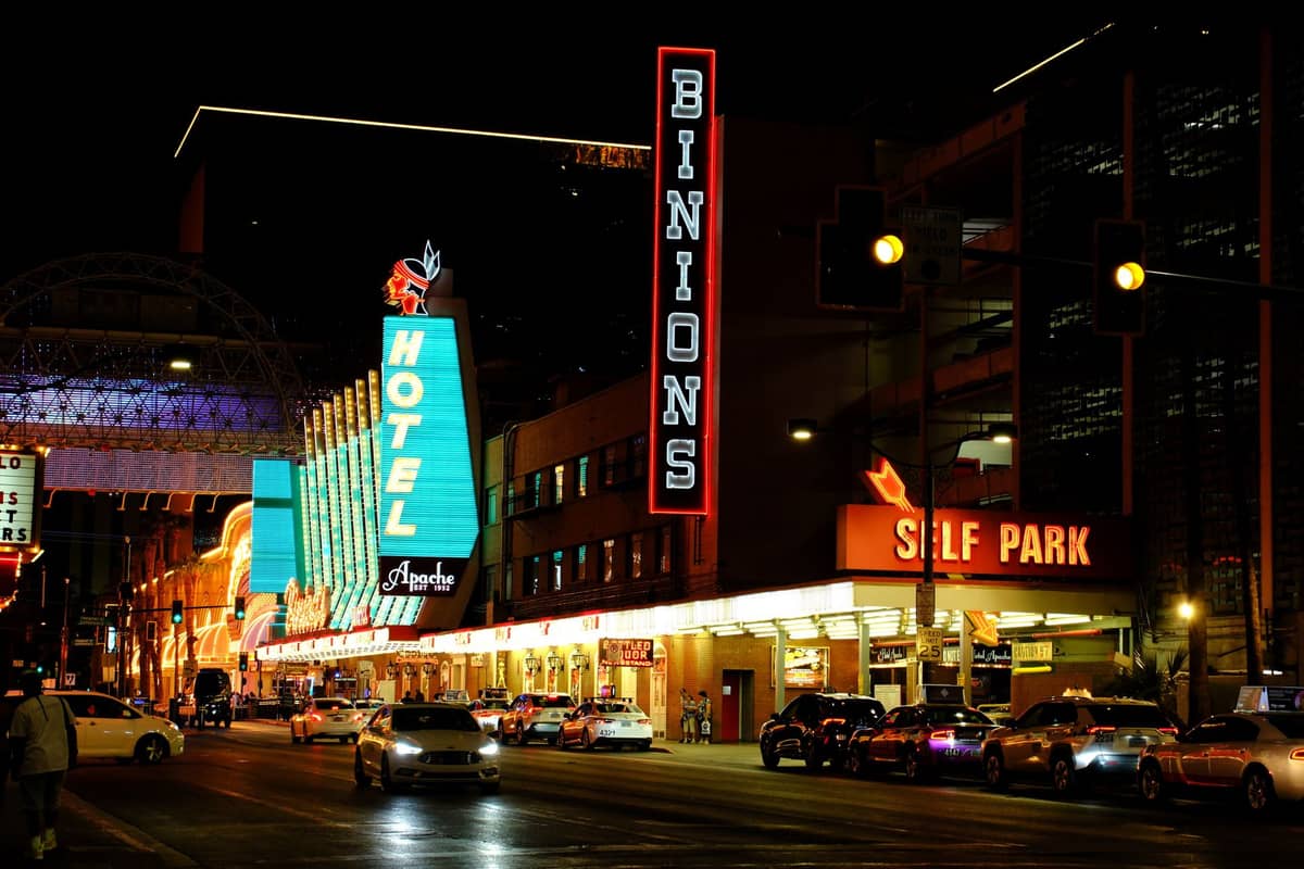 Landscape photo of Las Vegas' Fremont Street at night, featuring neon signs for Binion's and Hotel Apache, with cars and people enjoying the lively nightlife.