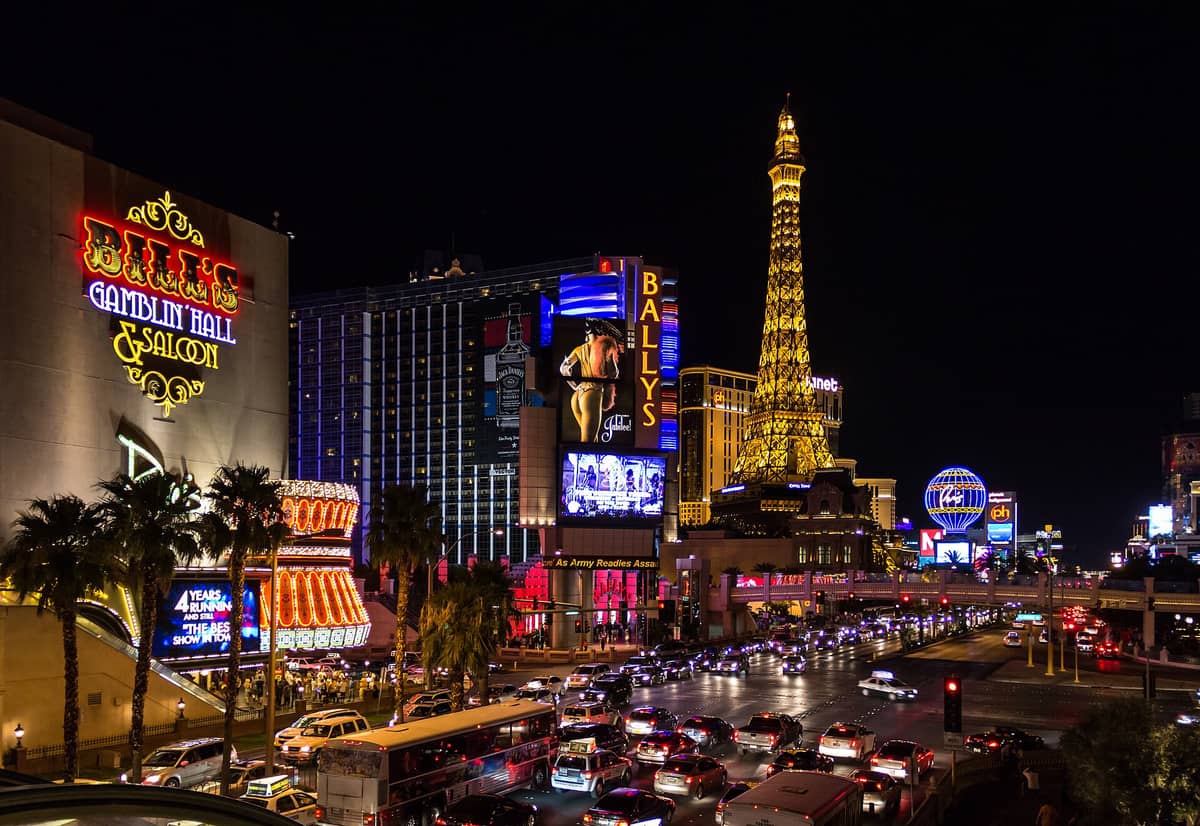 Night view of Las Vegas Strip with Bally's, the illuminated Eiffel Tower replica at Paris Las Vegas, neon signs, and busy traffic.