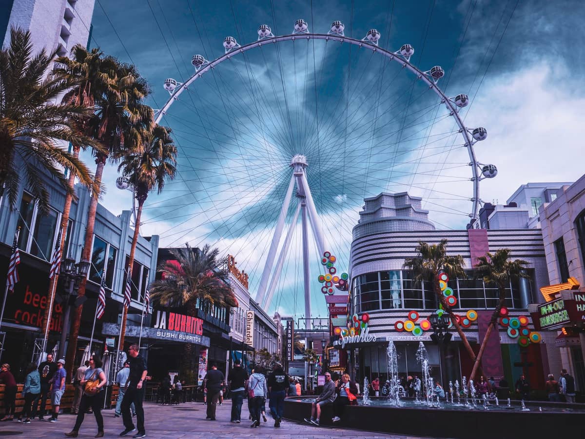The High Roller Ferris wheel at The LINQ Promenade in Las Vegas with people walking around, shops, and palm trees under a blue sky.