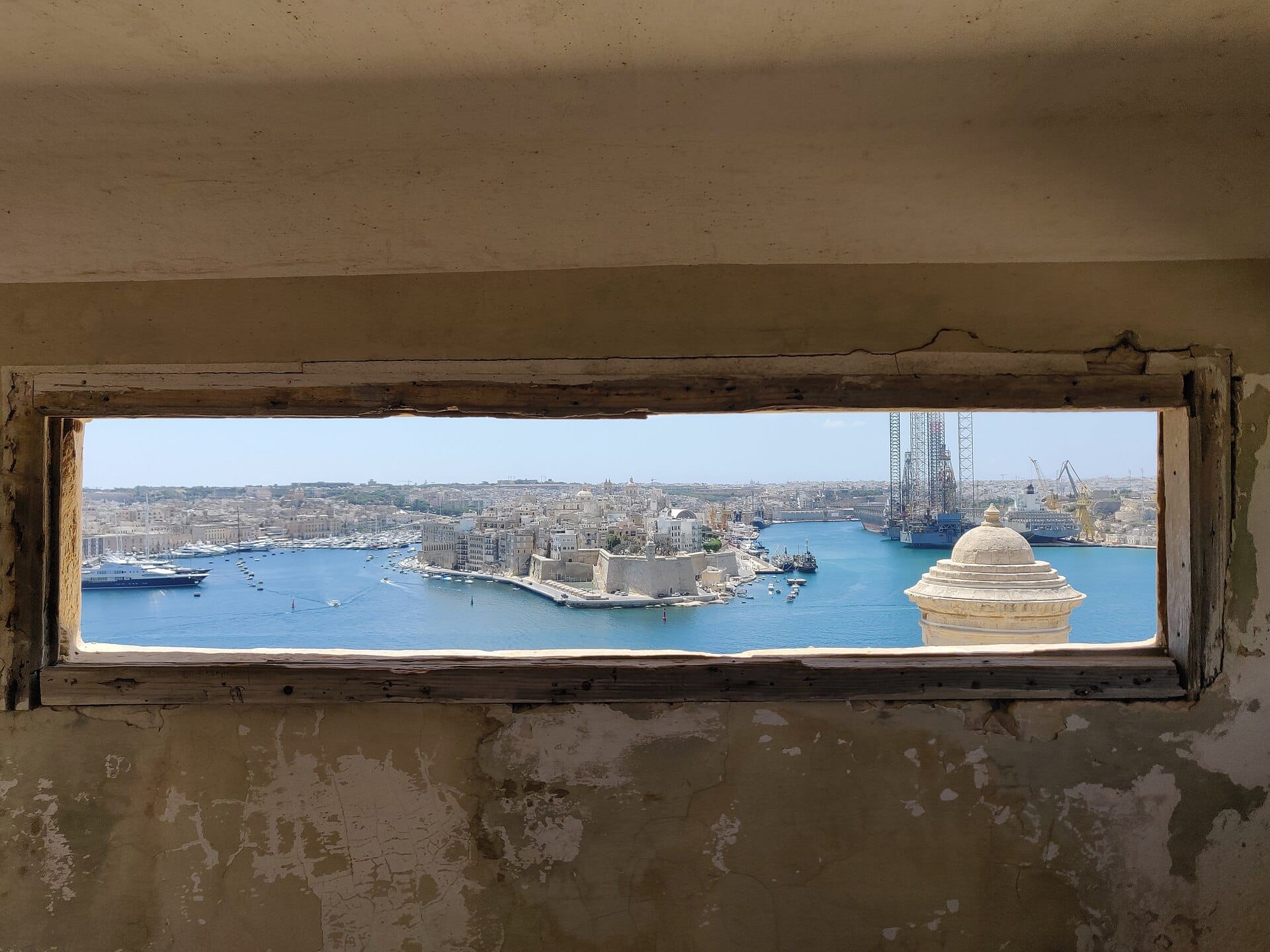 Vista del Grand Harbour di Valletta e del paesaggio urbano attraverso una finestra consumata all'interno delle Lascaris War Rooms, con storiche mura in pietra e mare azzurro.