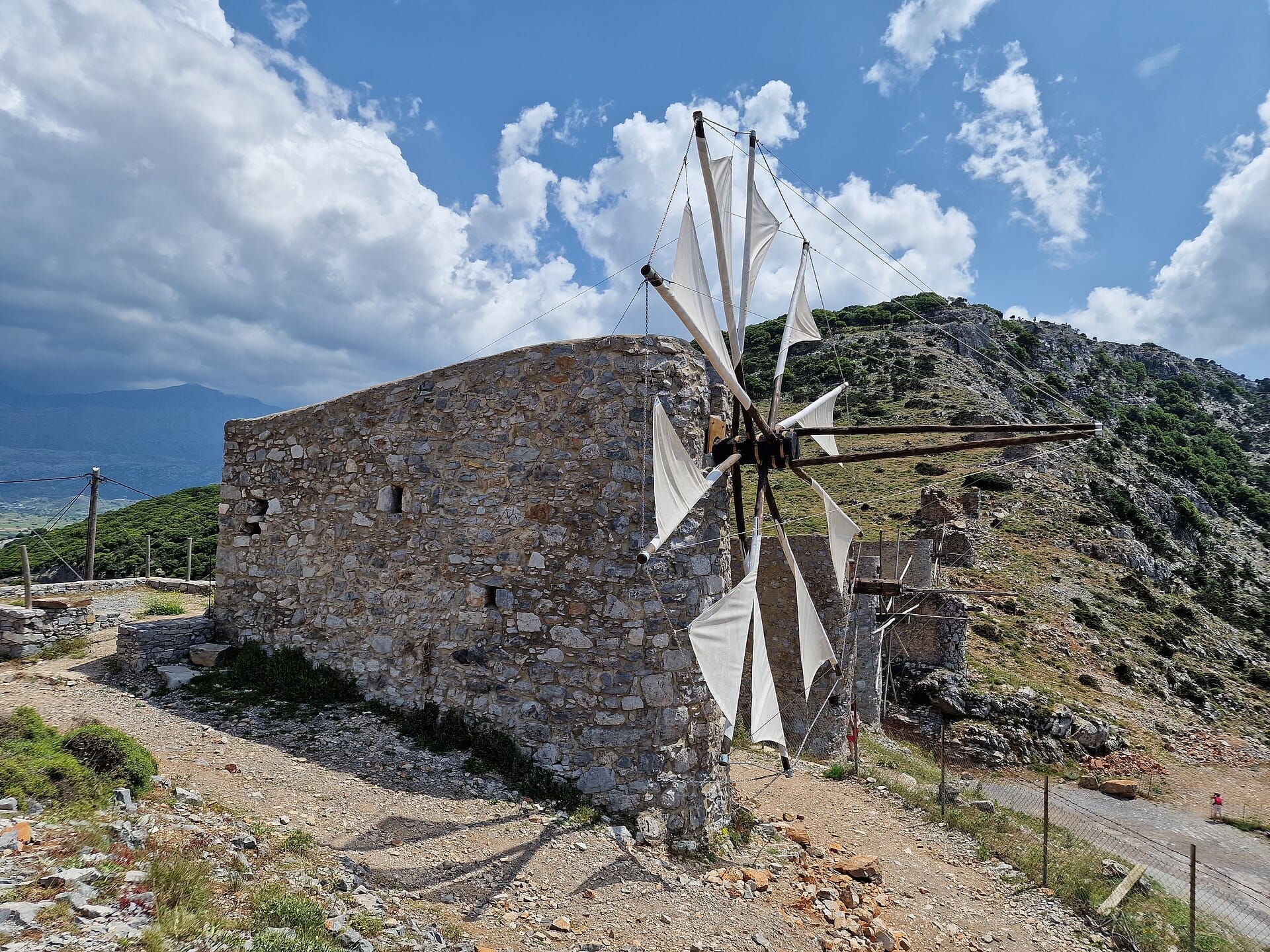 Mulino a vento in pietra con vele di tela bianca sull'Altopiano di Lassithi, con colline aspre sullo sfondo e un cielo luminoso e parzialmente nuvoloso a Creta.