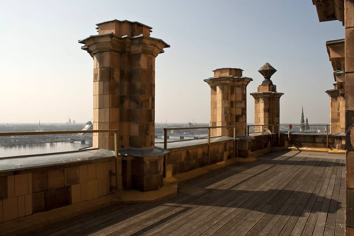 Steinpfeiler und Holzterrasse des Aussichtspunkts Panorama Riga mit Blick auf die Stadt, den Fluss und ferne Türme unter klarem Himmel.