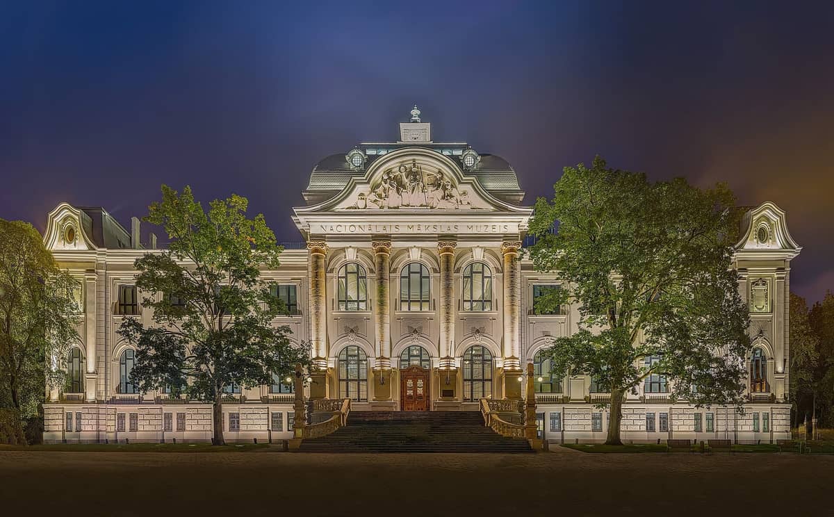Nächtliche Ansicht der beleuchteten neoklassizistischen Fassade des Lettischen Nationalen Kunstmuseums in Riga, mit imposanten Säulen, aufwendigen Details und Bäumen, die den Eingang rahmen.