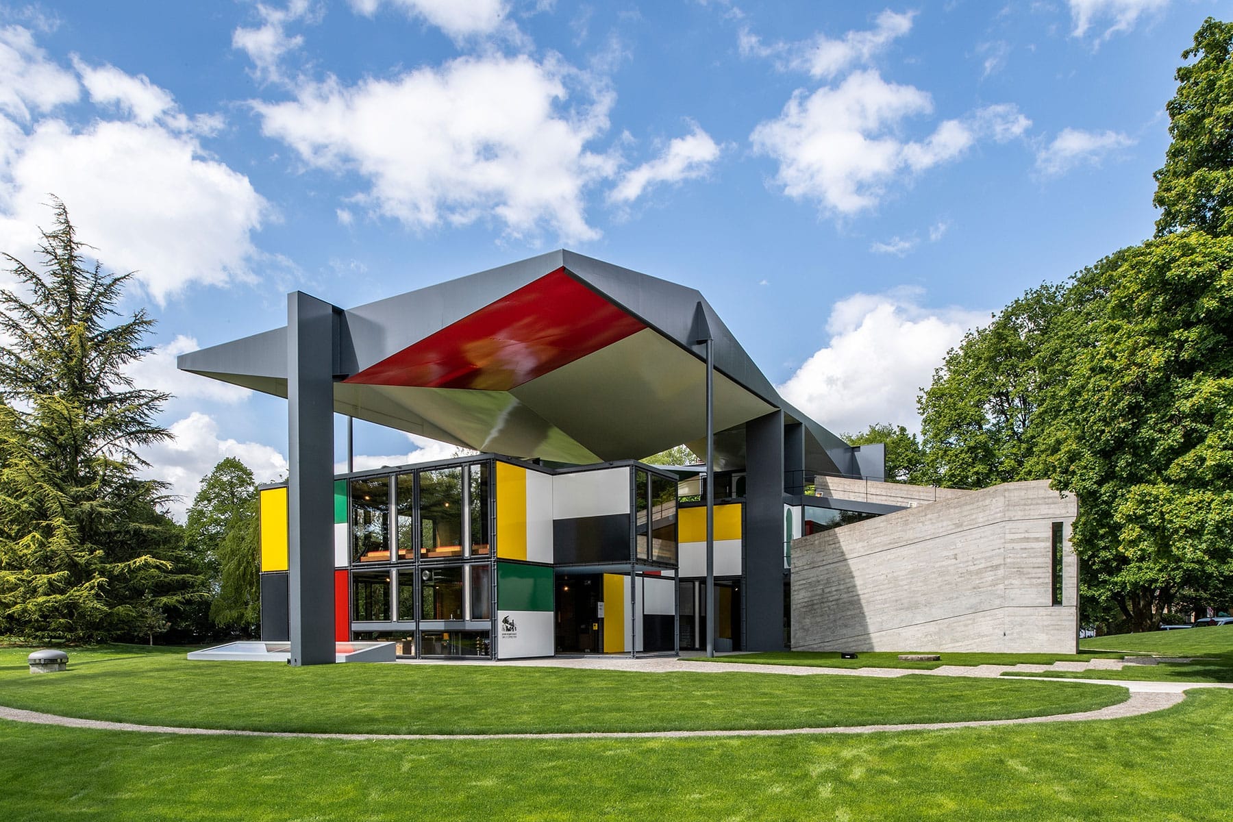 Pavillon Le Corbusier ในซูริก: อาคารโมเดิร์นนิสต์ที่ทำจากเหล็ก กระจก และแผงสีสันสดใส ล้อมรอบด้วยสนามหญ้าสีเขียวและท้องฟ้าสีฟ้า