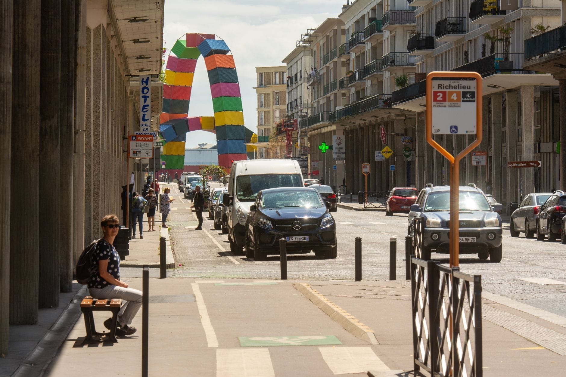 Scena uliczna w Le Havre przedstawiająca przystanek autobusowy, nowoczesną architekturę, pieszych, ruch uliczny i kolorową bramę instalacji artystycznej.