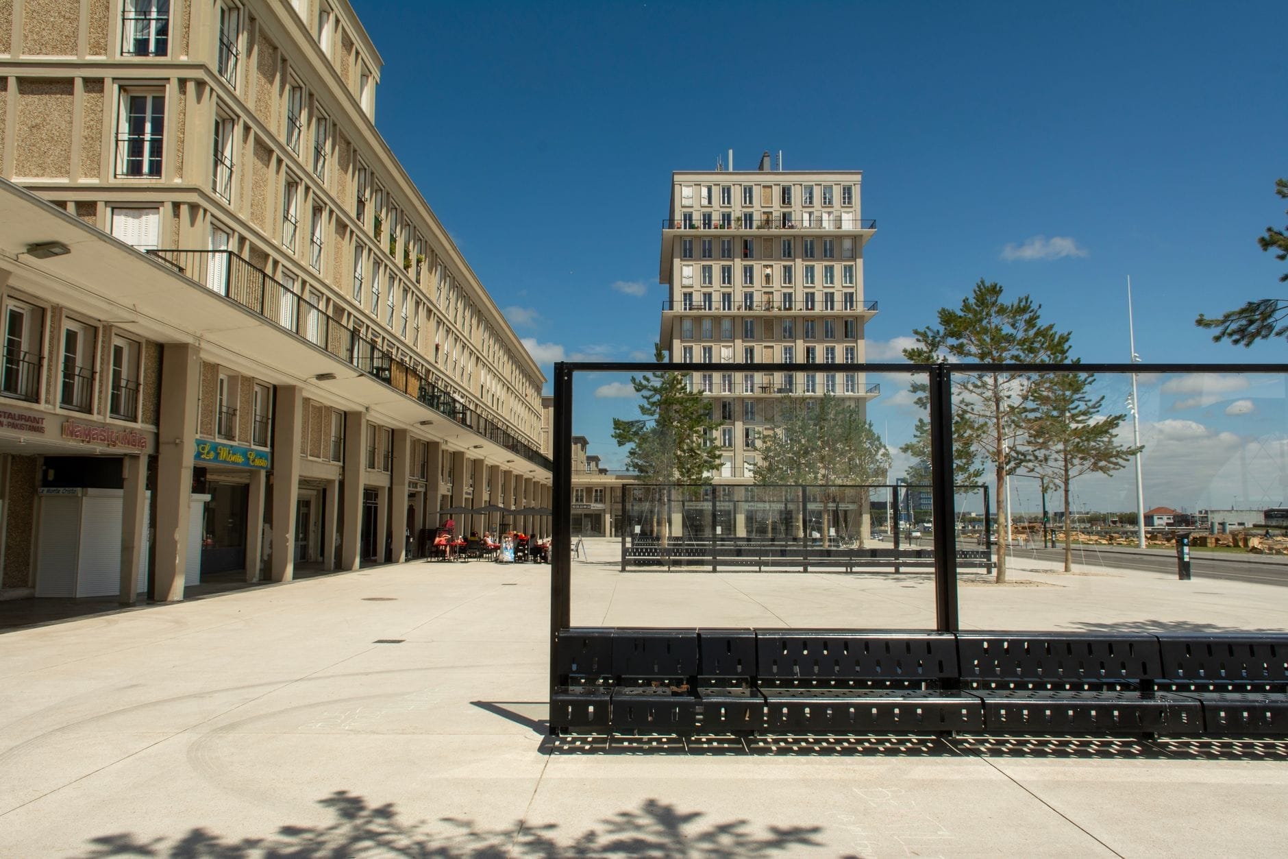 Le Havre'da modern bir meydan, belirgin yüksek bir bina, ağaçlar ve boyalı bir manzarayı çağrıştıran boş siyah metal çerçeve.