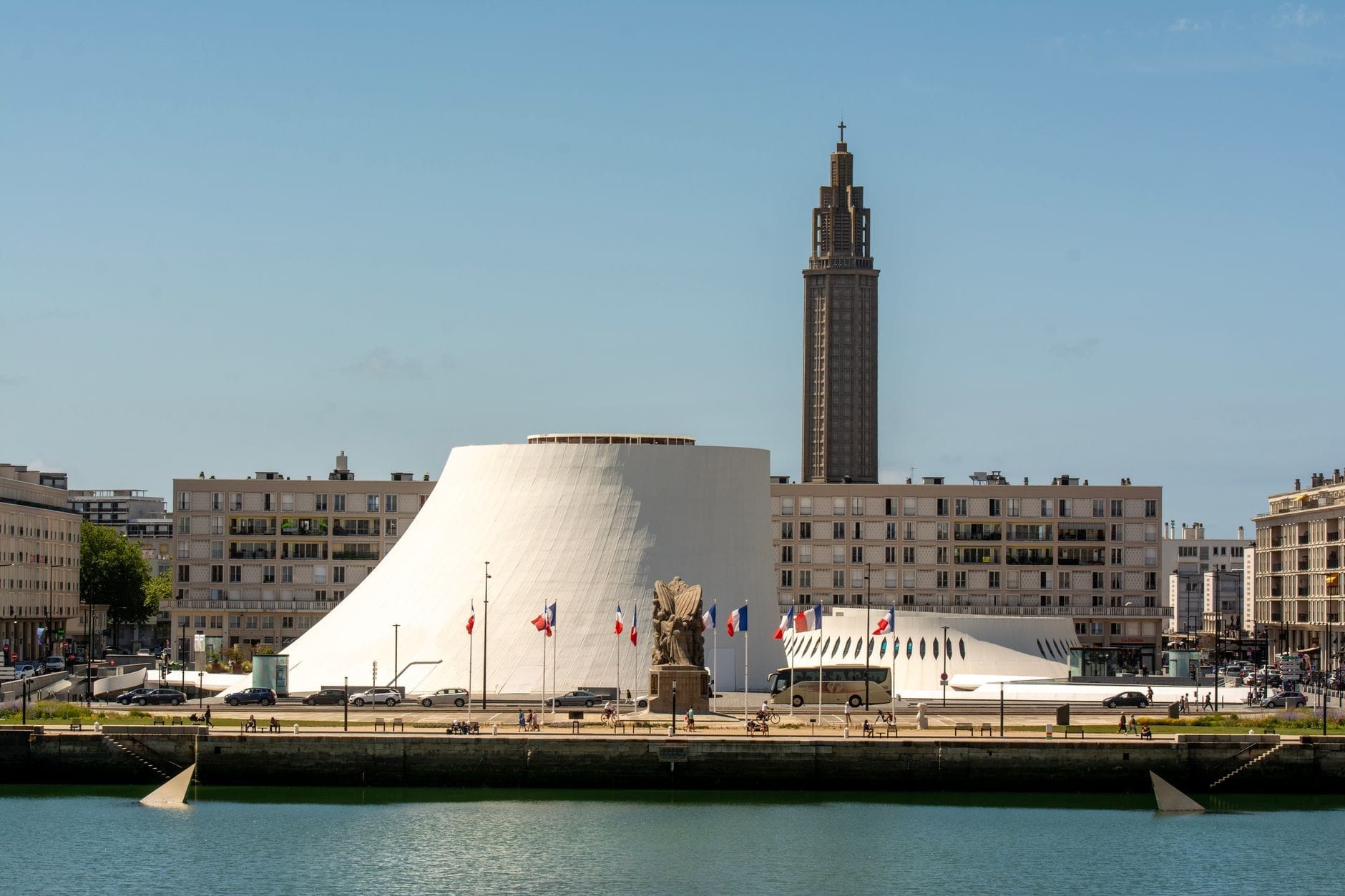 Vista desde el agua hacia Le Volcan, el icónico centro cultural modernista y espacio de exposiciones de Le Havre, con el paisaje urbano circundante y la torre de la iglesia St. Joseph al fondo.