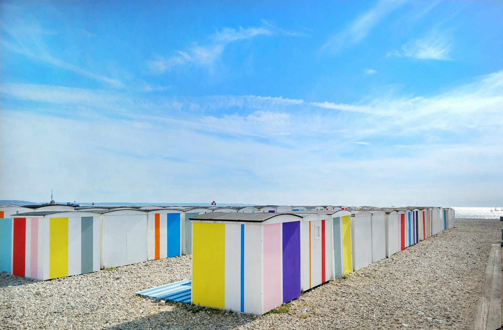 Capanne da spiaggia colorate su una riva di ciottoli a Le Havre sotto un cielo estivo azzurro, con il mare e la spiaggia aperta sullo sfondo.