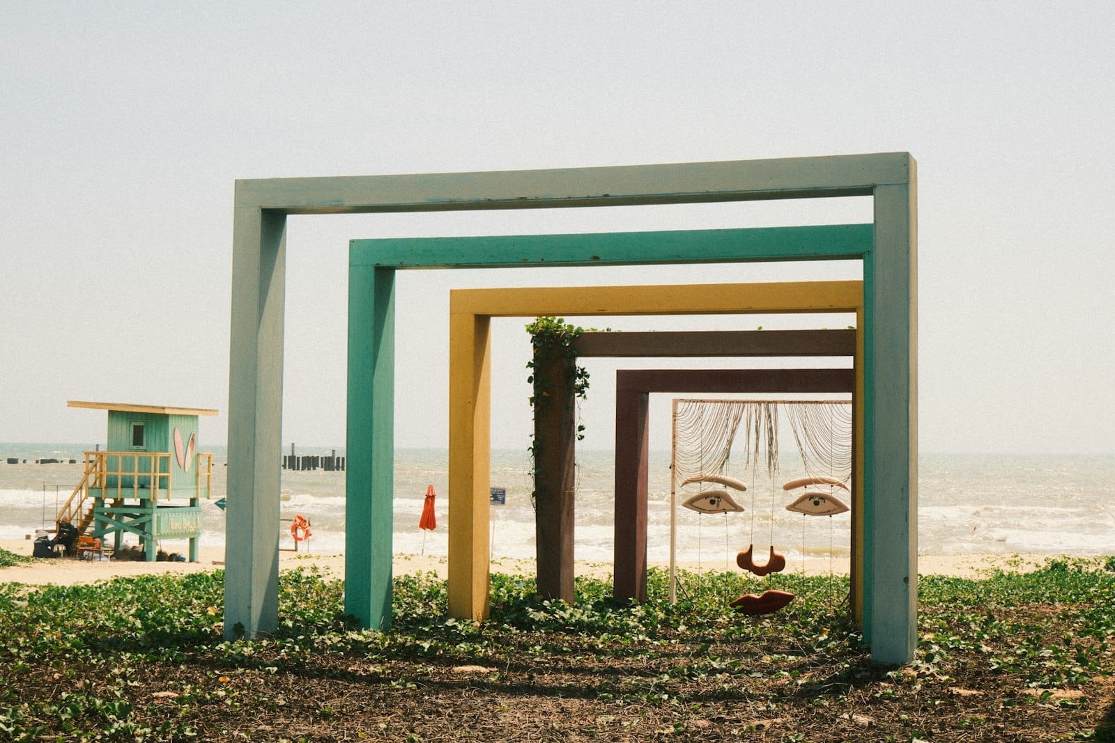 Marcos rectangulares de colores y arte lúdico con rostros instalados al aire libre sobre hierba cerca del paseo marítimo, con olas y una playa al fondo.