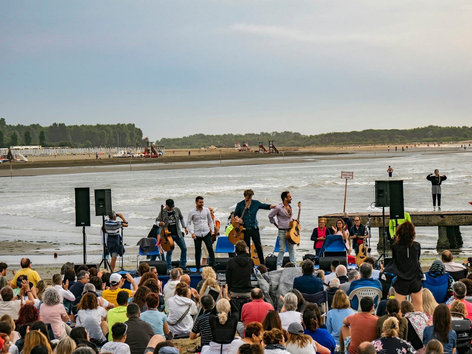 Concierto al aire libre en una playa con una multitud sentada viendo a músicos actuar cerca de la orilla del agua.