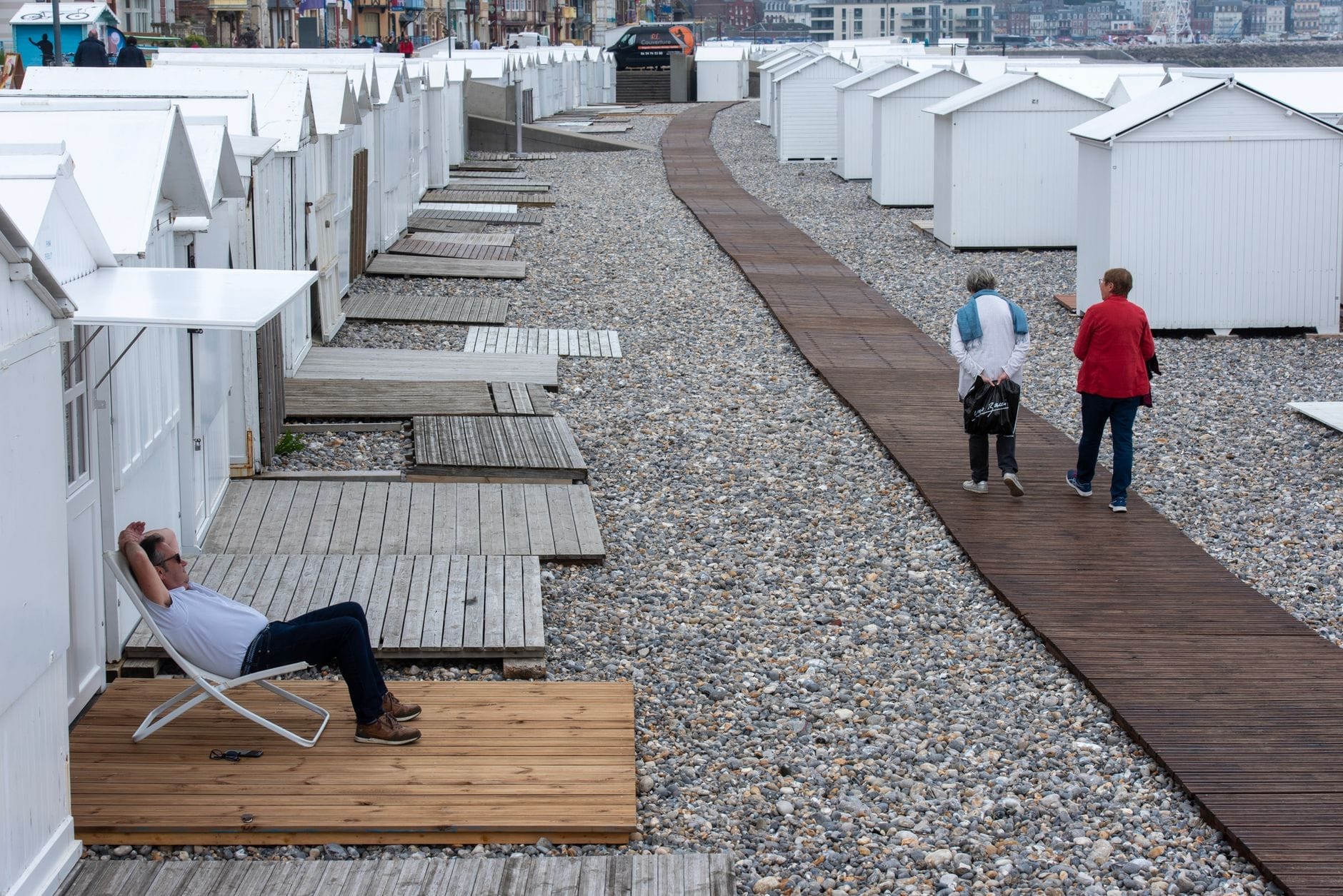 Playa de guijarros en Le Havre bordeada de casetas de playa blancas, pasarelas de madera y personas paseando o relajándose junto al paseo marítimo.