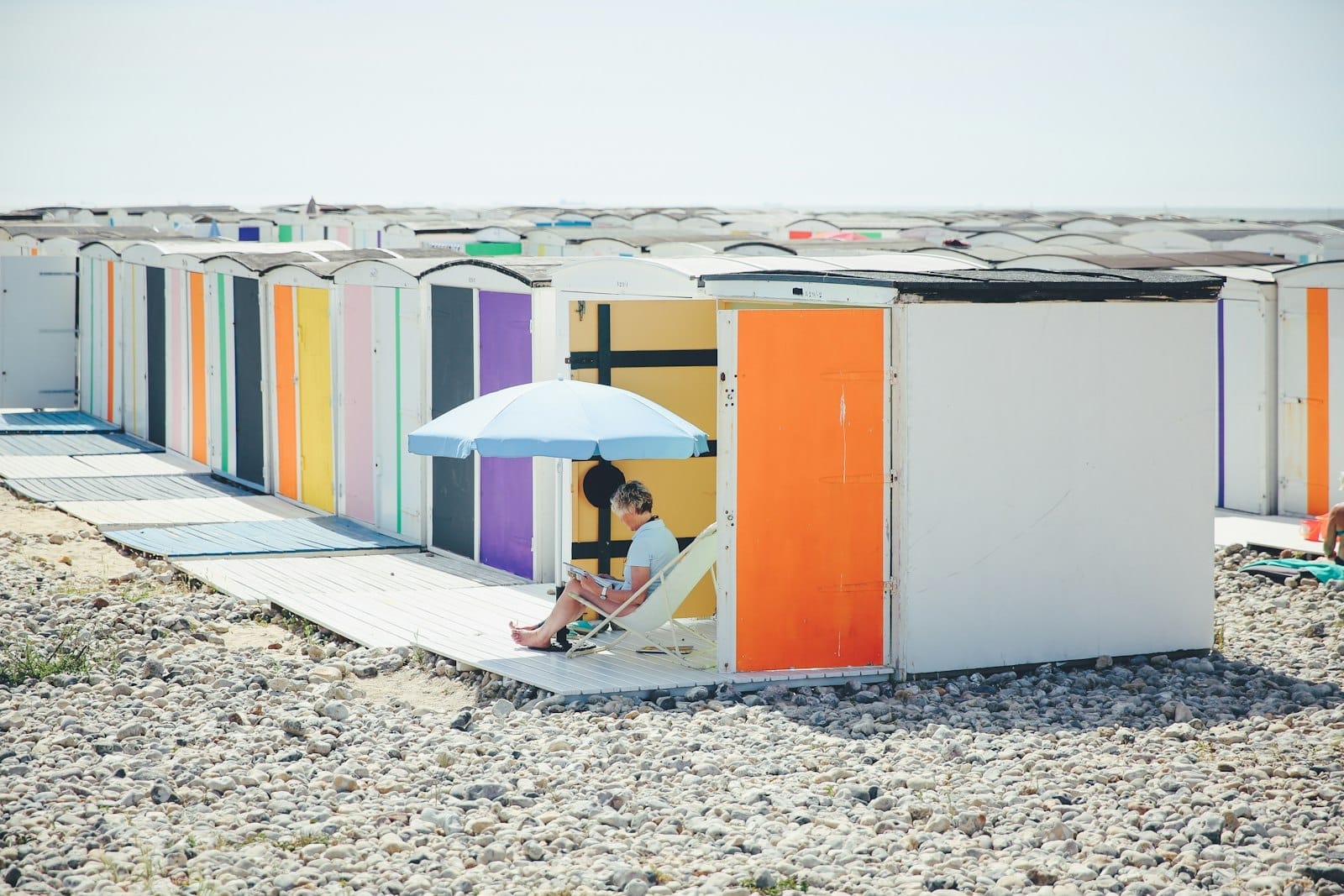 Cabines de praia coloridas ao longo da orla de seixos de Le Havre com uma pessoa relaxando sob um guarda-sol, evocando atividades de praia ao ar livre para toda a família.