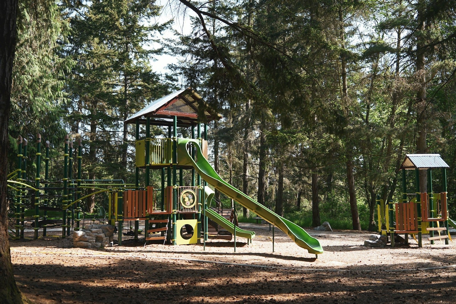 Grande área de lazer verde rodeada de árvores altas com escorregadores e estruturas de escalada em um parque arborizado.