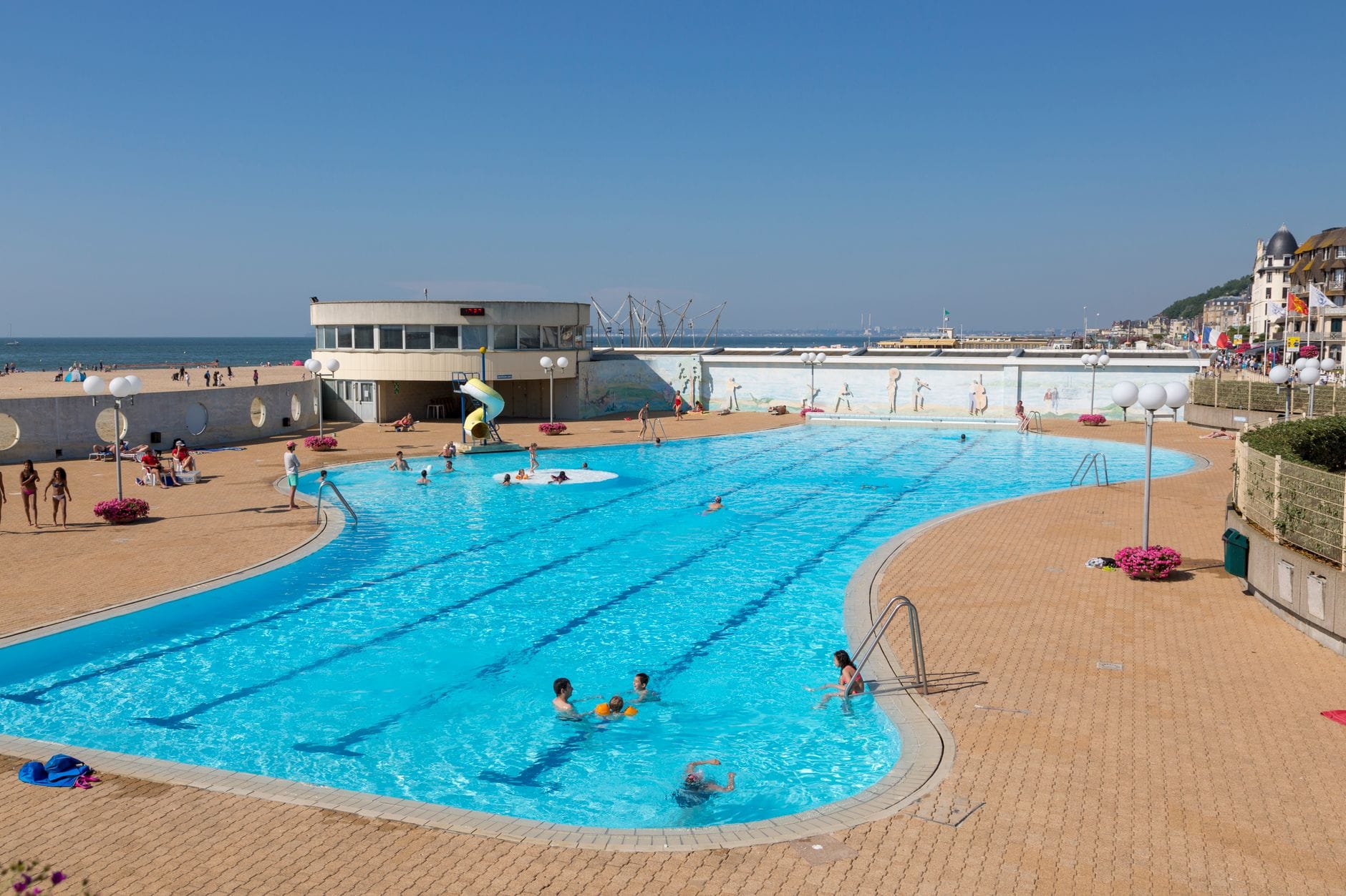 Piscina all'aperto accanto a una spiaggia sabbiosa con bambini e famiglie che nuotano, cielo blu e il vivace lungomare di Le Havre sullo sfondo.