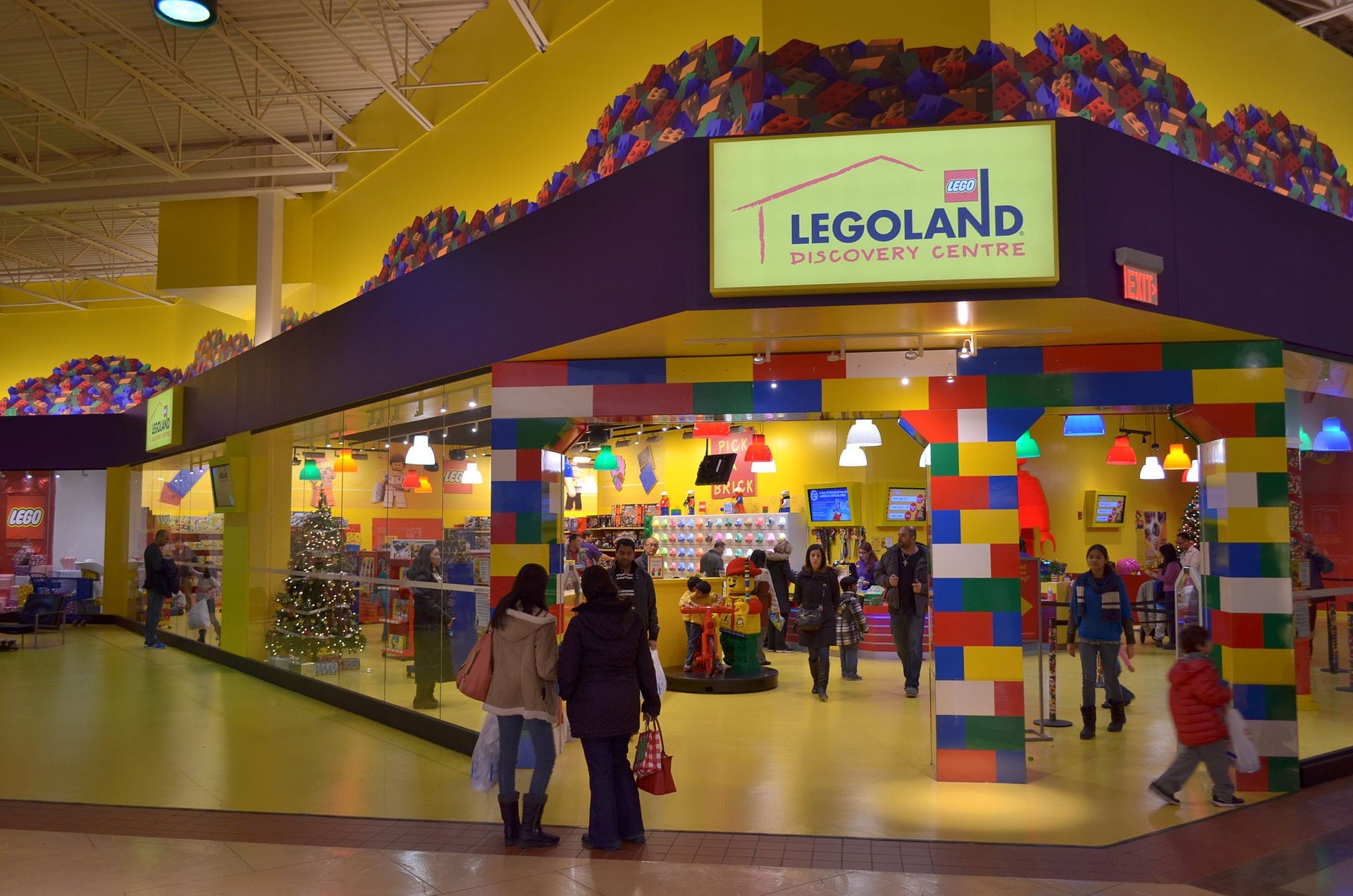 ทางเข้า LEGOLAND Discovery Centre Toronto ที่ Vaughan Mills พร้อมซุ้มประตูอิฐ LEGO หลากสีสัน กำแพงสีเหลืองสดใส และครอบครัวที่กำลังเข้าชมสถานที่ท่องเที่ยว