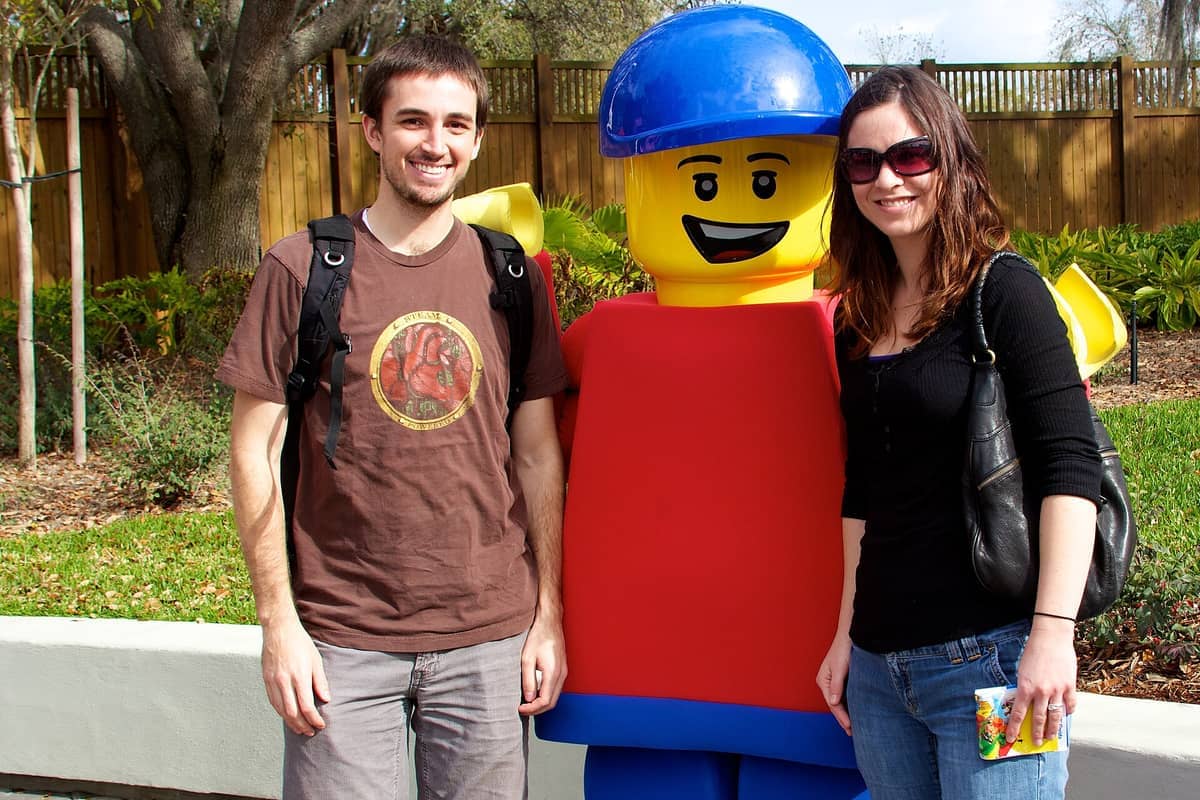 Zwei lächelnde Besucher posieren mit einer großen Lego-Figurenstatue im LEGOLAND Florida Resort, umgeben von Grünpflanzen und einem Holzzaun an einem sonnigen Tag.