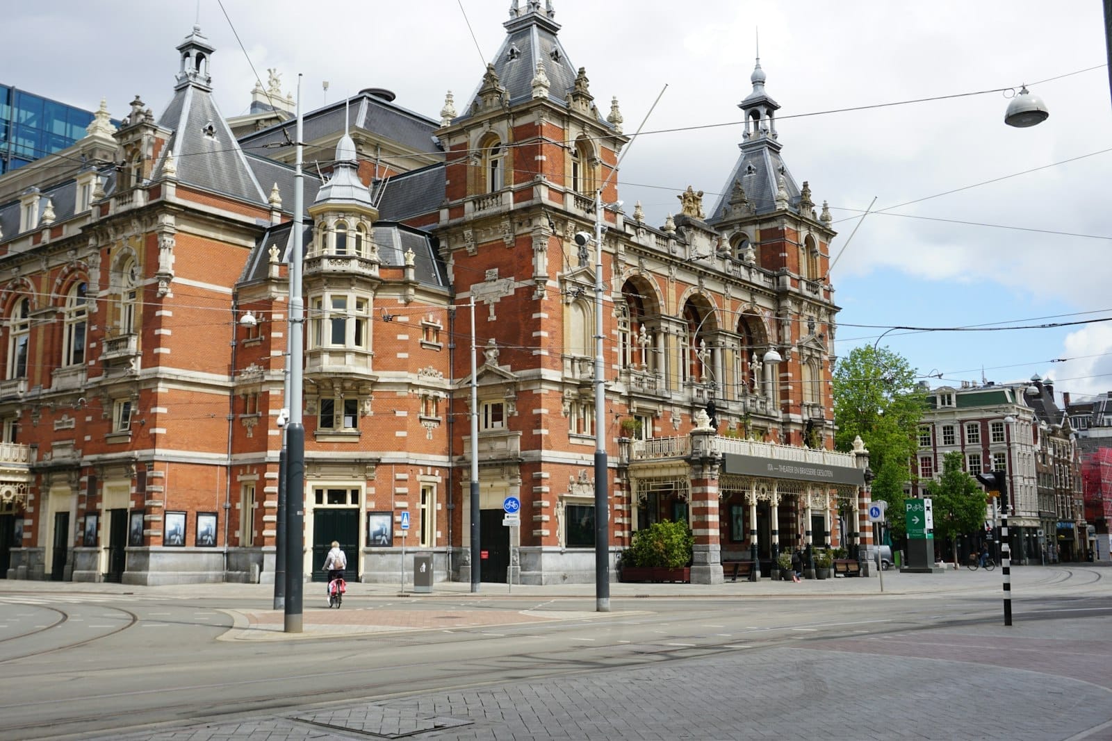 Teatro Stadsschouwburg en el Leidseplein de Ámsterdam, con su ornamentada arquitectura de ladrillo rojo, vías de tranvía y un ciclista pasando en un día nublado.