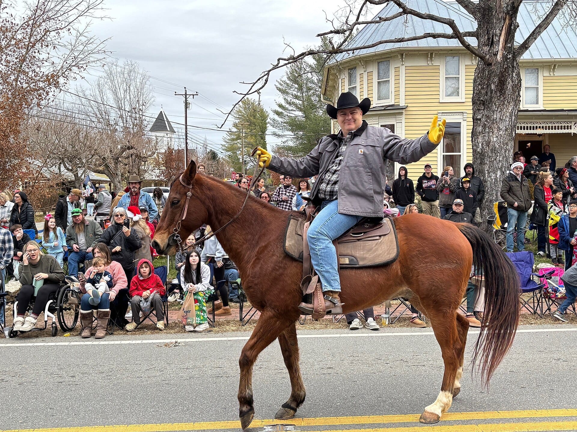 Un vaquero a caballo saluda a la multitud durante un desfile en Leiper's Fork Village, con casas históricas y árboles de invierno al fondo.