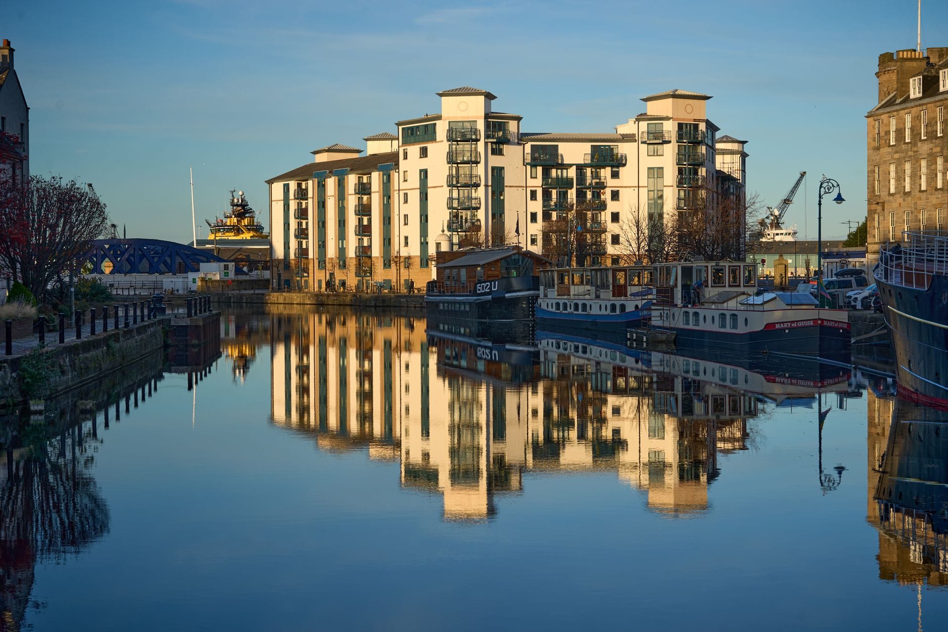 Das Leith-Ufer mit modernen Apartments, angedockten Booten und dem ruhigen Wasser des Hafenviertels, das Gebäude und blauen Himmel spiegelt.