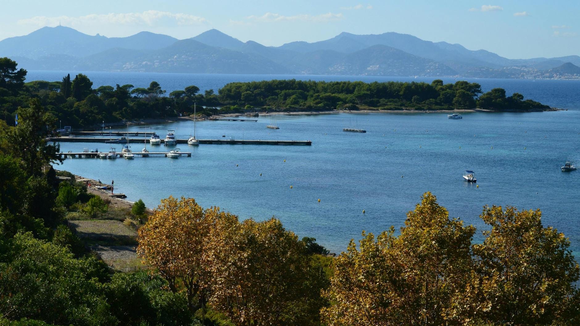 Vista panorâmica das Ilhas Lérins com barcos ancorados na marina, pinheiros exuberantes e montanhas ao fundo, convidando os visitantes a explorar a natureza perto de Cannes.