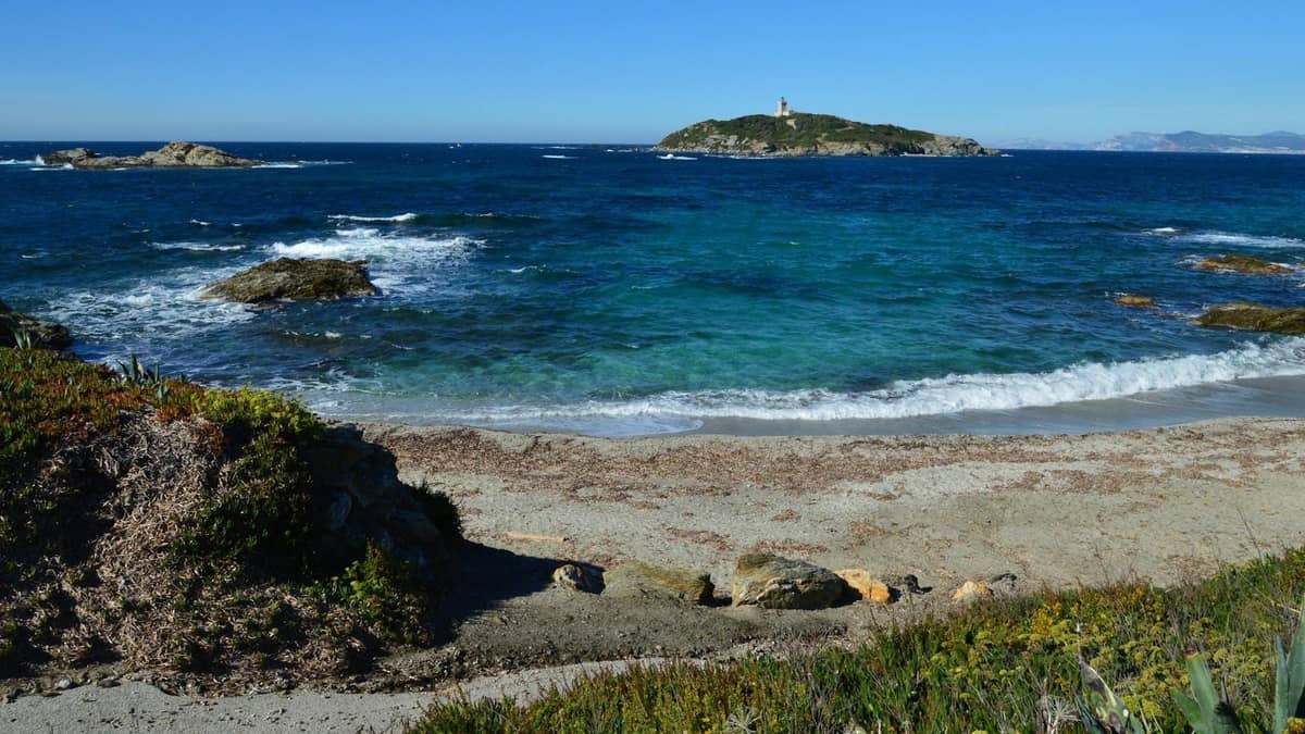 Blick auf eine kleine mediterrane Insel mit einem Leuchtturm, über blaues Wasser von einem natürlichen Sandstrand und einer felsigen Küste aus gesehen.