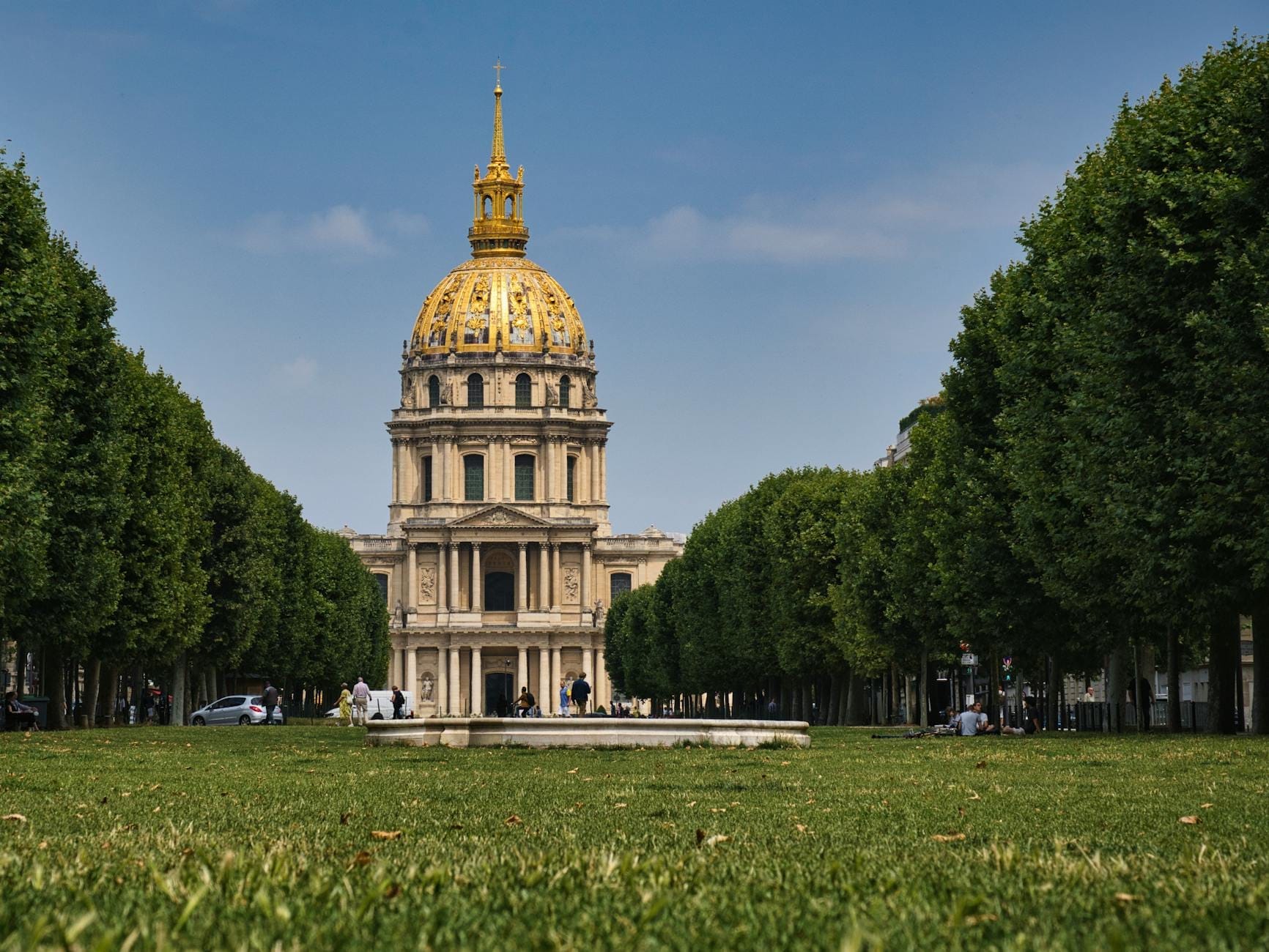 Vista frontale della cupola dorata di Les Invalides a Parigi, incorniciata da un prato verde e alberi frondosi sotto un cielo azzurro luminoso.