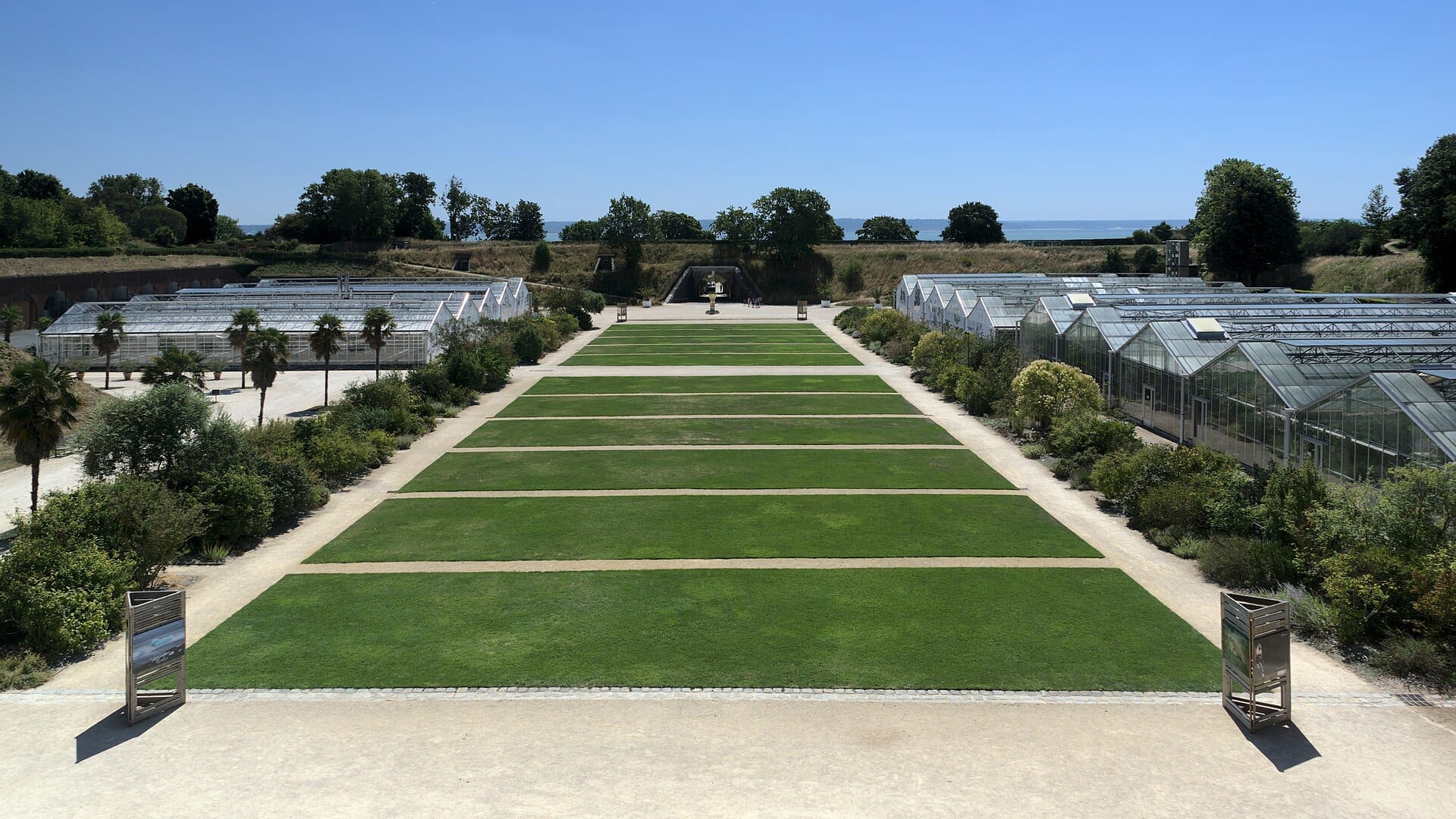 Prato centrale ai Les Jardins Suspendus a Le Havre, fiancheggiato da serre e rigogliose bordure sotto un cielo azzurro sereno.