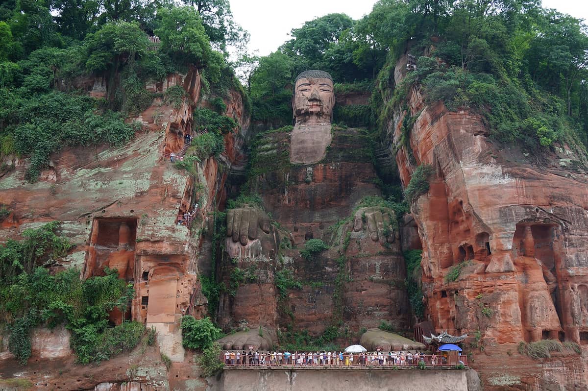 Malawakang tanawin ng Leshan Giant Buddha na inukit sa pulang sandstone na bangin, napapaligiran ng luntiang mga puno, may mga turistang nakatayo sa paanan ng rebulto.