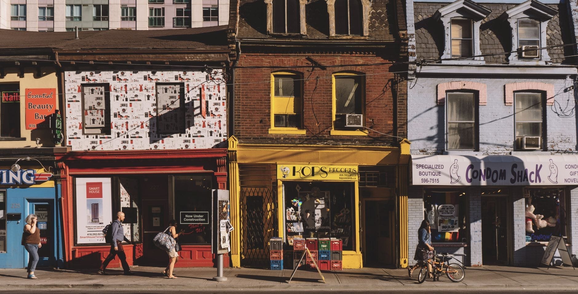 ร้านค้าหลากสีสันรวมถึง Kops Records และ Condom Shack บน Queen Street East ใน Leslieville, Toronto พร้อมผู้คนที่กำลังเดินและขี่จักรยานใต้แสงแดดยามบ่าย