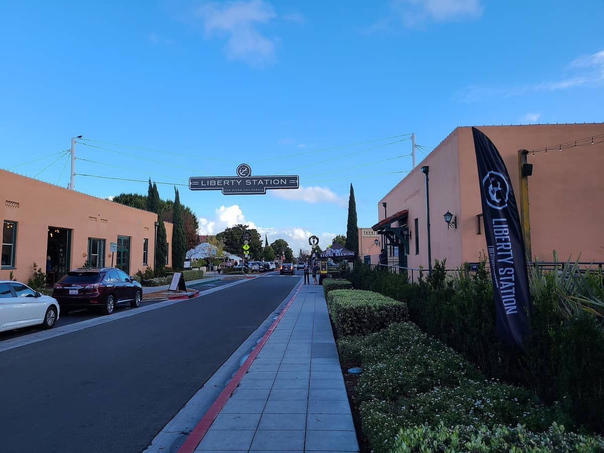 Eingang der Liberty Station in San Diego, mit pfirsichfarbenen Gebäuden, geparkten Autos, grünen Büschen und einem klaren blauen Himmel im Hintergrund.