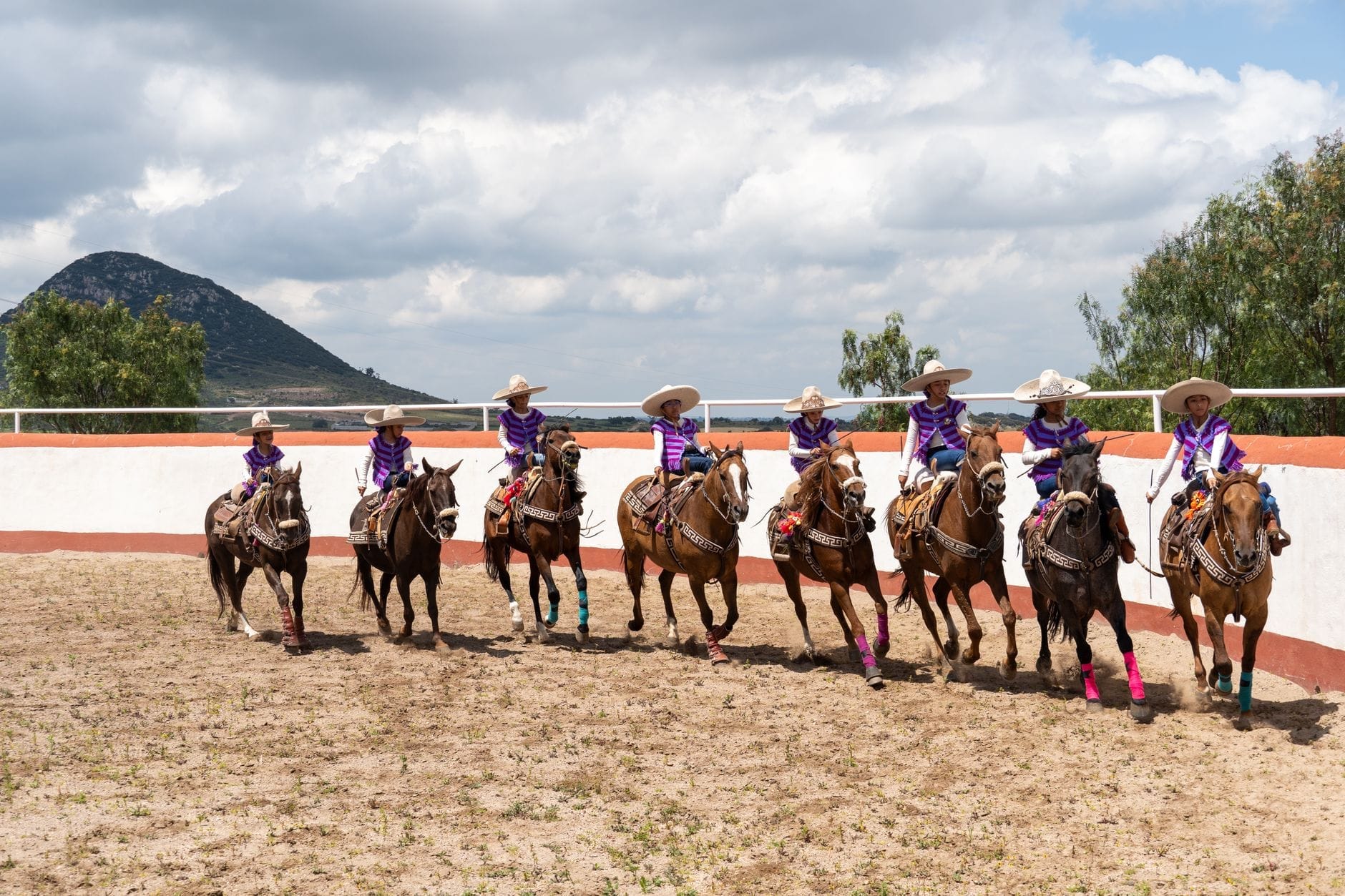 Otto escaramuzas in abiti colorati coordinati e grandi sombreros cavalcano in formazione all'interno di una tradizionale arena charro, con le montagne e un cielo azzurro sullo sfondo.