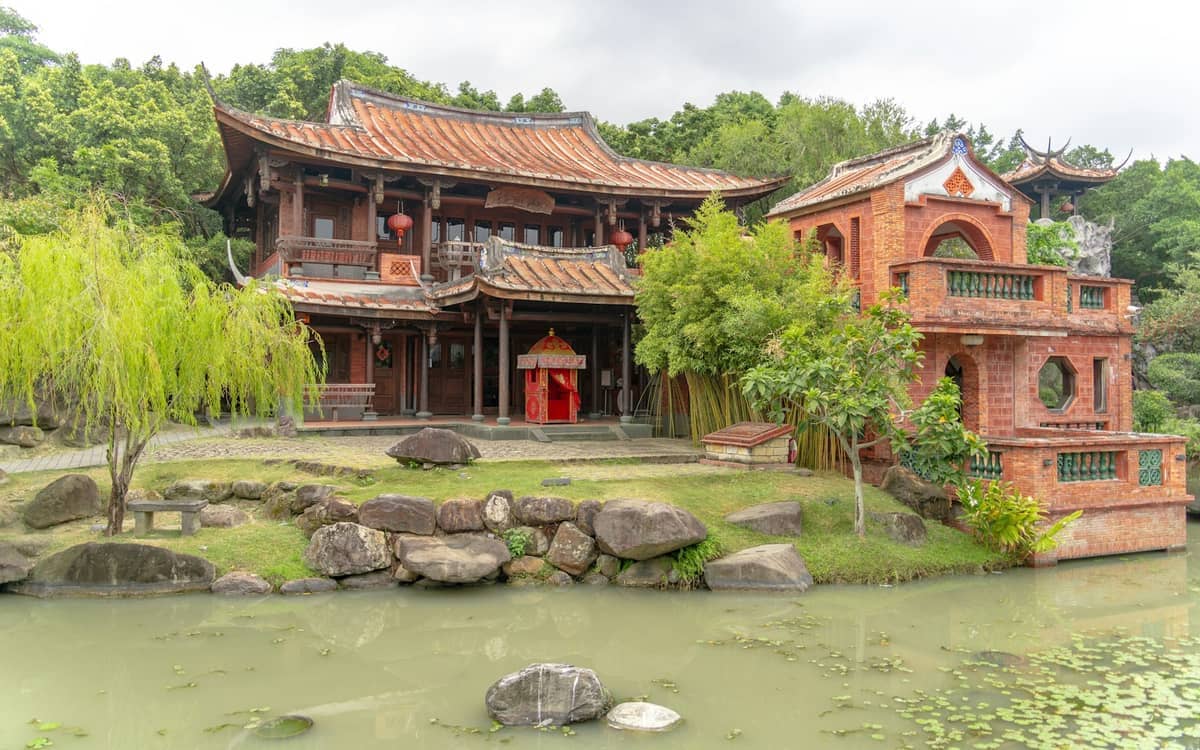 Das historische Haus Lin An Tai mit seiner traditionellen Holzarchitektur im Minnan-Stil, roten Laternen, üppigen grünen Gärten und einem ruhigen Teich unter bewölktem Himmel.