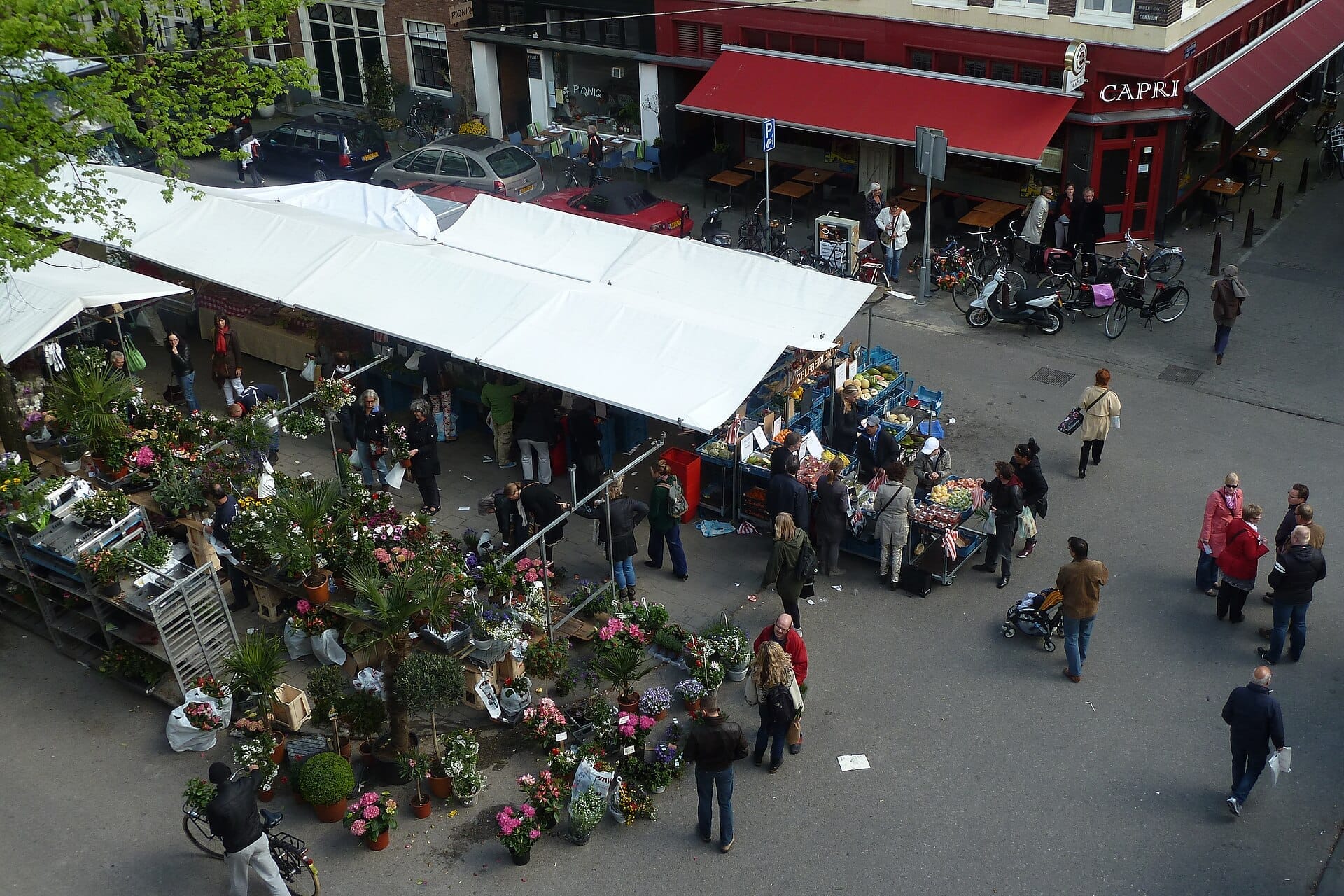 Vista aérea del mercado Lindengracht con puestos de flores, verduras y personas paseando bajo toldos blancos en una concurrida esquina.