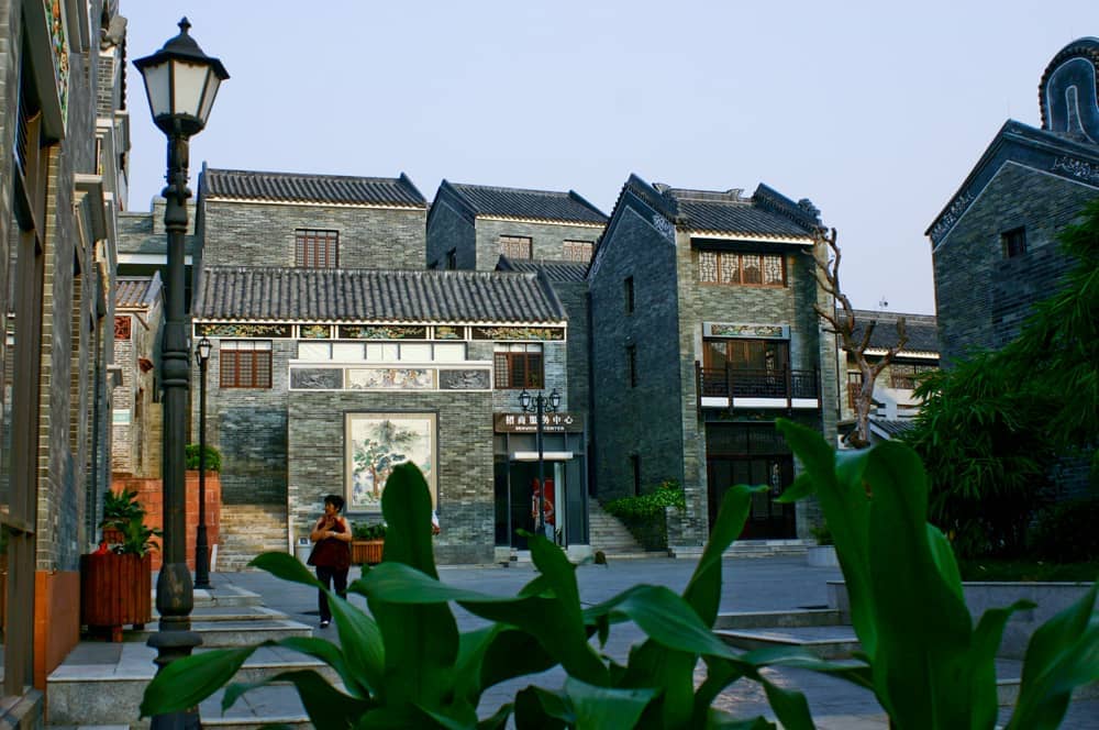 Traditional gray brick buildings at Lingnan Impression Park with intricate woodwork, lush greenery, and a lone visitor walking in the courtyard.