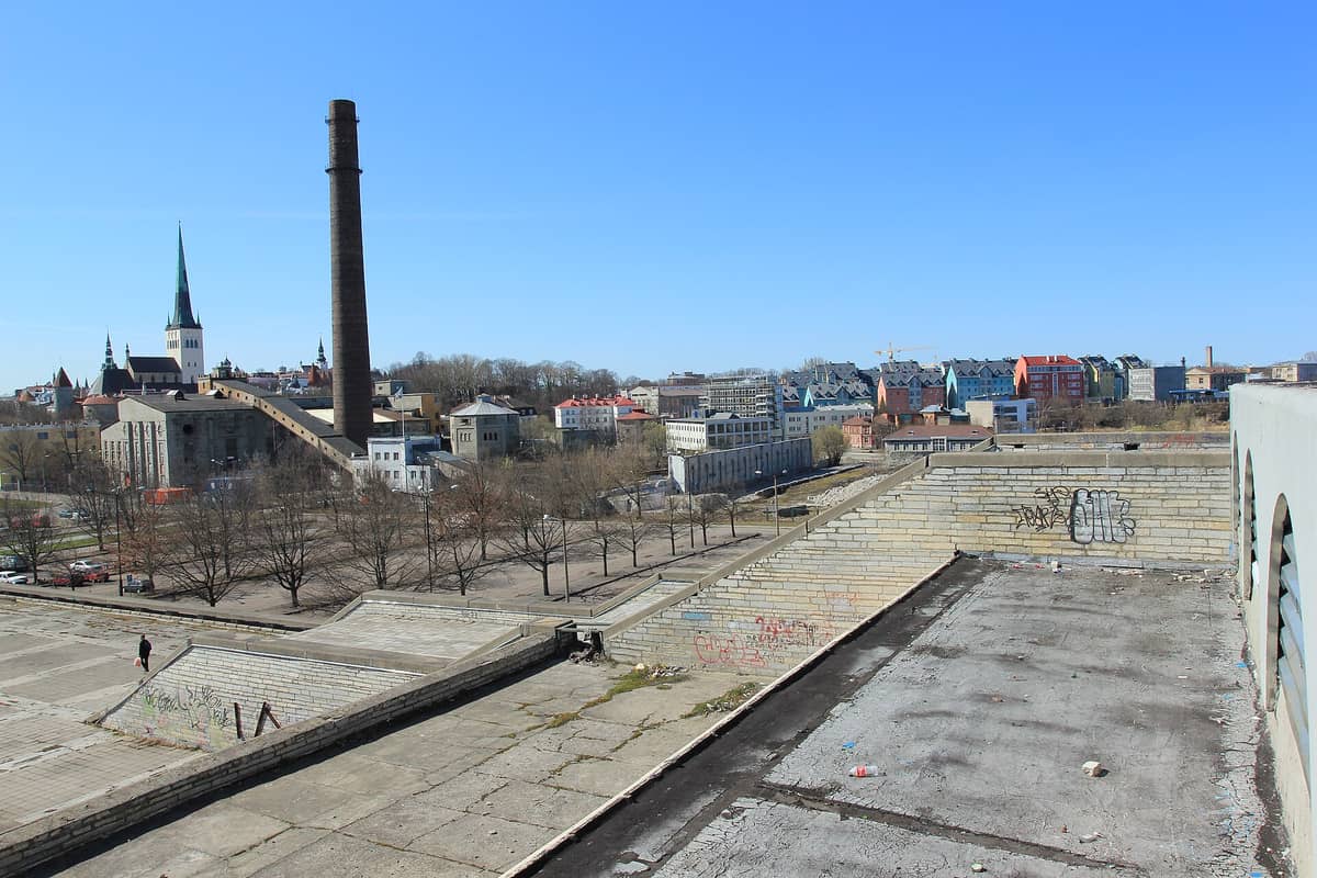 Blick vom Dach des Linnahall auf seine verfallenden Betonterrassen, Graffiti und das Panorama Richtung Tallinns Altstadt und den hohen Backsteinschornstein.