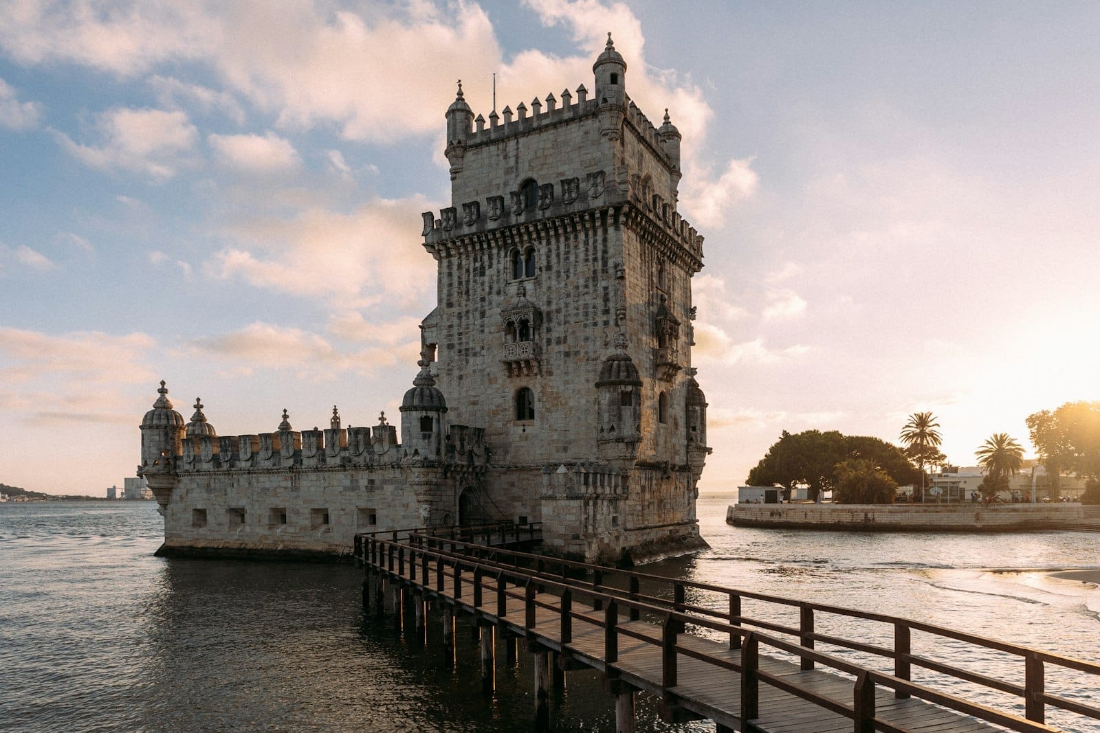 Torre de Belém em Lisboa ao pôr do sol, vista de uma passarela de madeira com suave luz dourada e detalhes nítidos do histórico monumento à beira do rio.