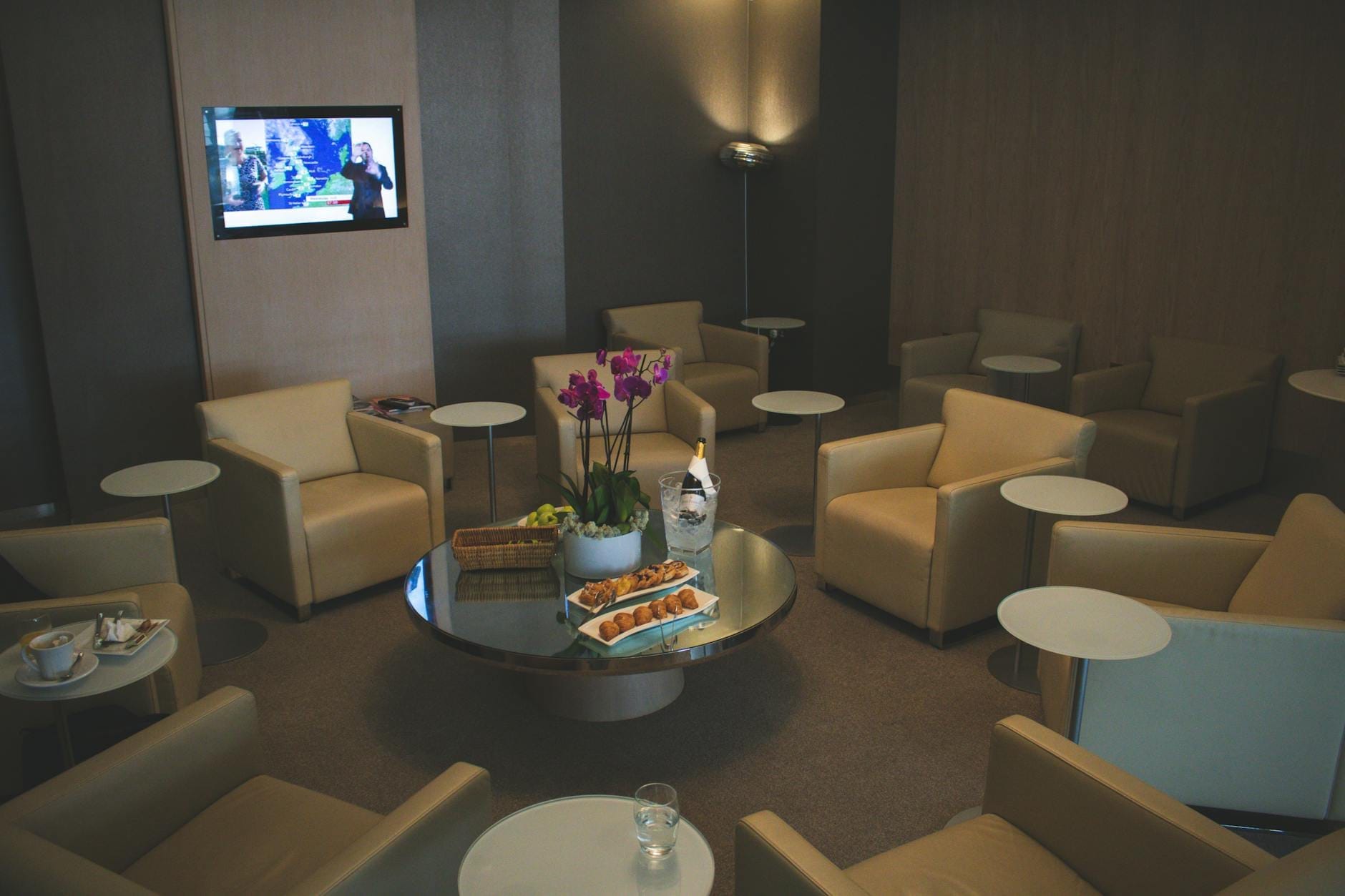 Sala VIP aconchegante do aeroporto com poltronas, mesas redondas, petiscos e flores em uma mesa central, e uma TV na parede.