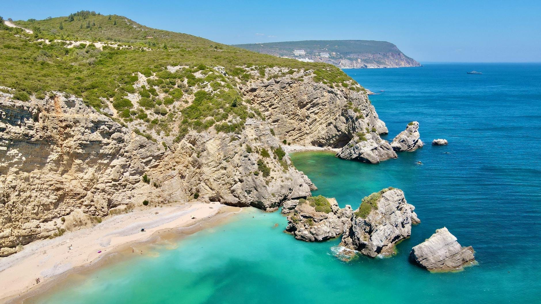 Falésias de calcário e água turquesa com uma praia de areia na Arrábida perto de Lisboa, vistas num dia claro e ensolarado.
