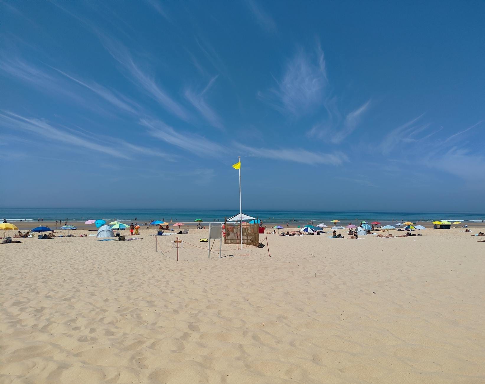 Ampla cena de praia de areia com guarda-sóis coloridos e banhistas sob um céu azul limpo, de frente para o Atlântico perto de Lisboa.